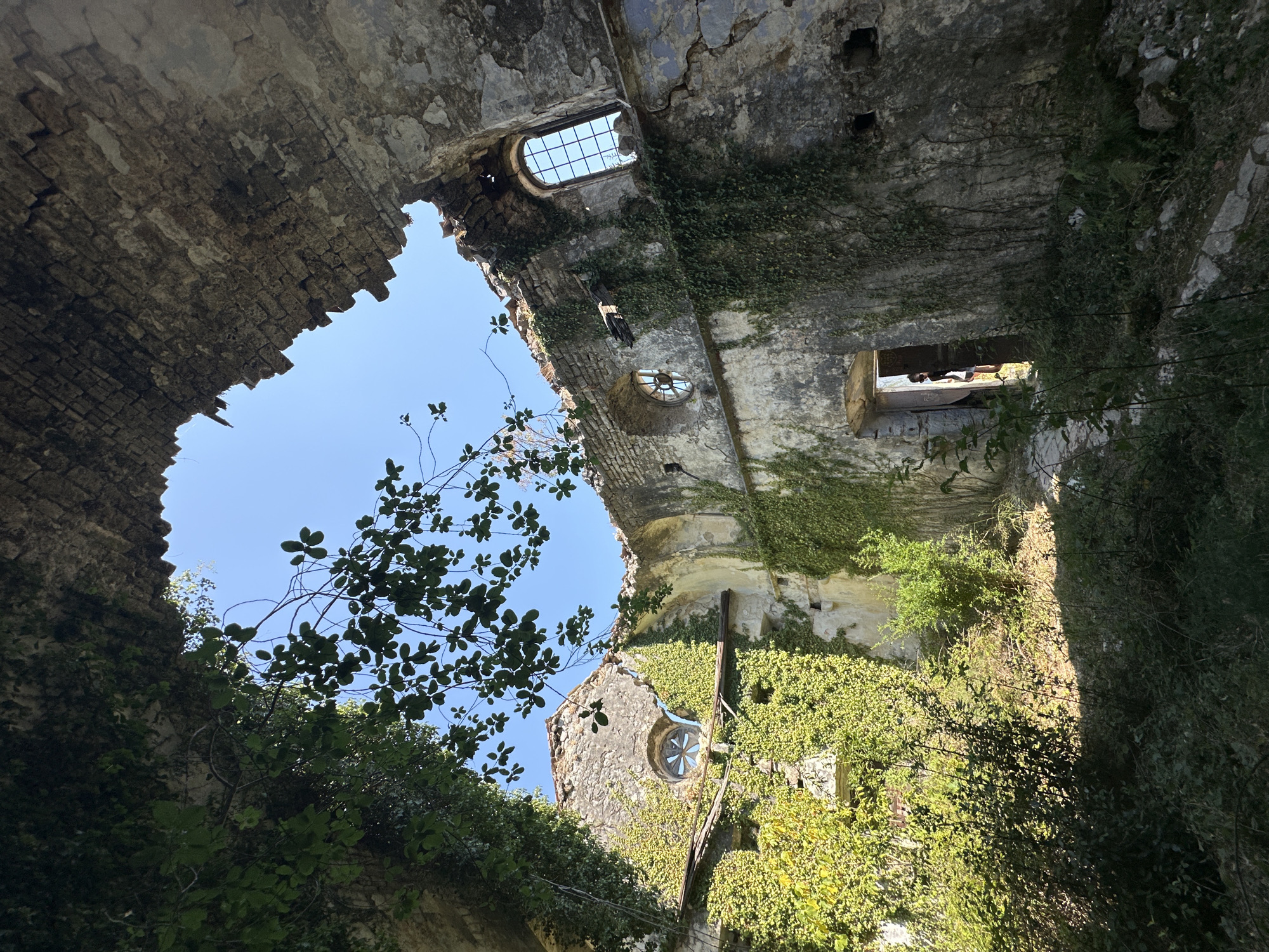 An interior view of a dilapidated stone building, likely a church or monastery, in Prčanj, Montenegro, is captured. The structure is heavily overgrown with vibrant green ivy, clinging to the crumbling walls and partially obscuring arched openings and windows. The roof is mostly gone, allowing sunlight to stream through an open section and illuminate the interior. One circular window with a star-shaped pane is visible on the left, while a larger rectangular window with a metal grate is on the right. In the center of the frame, a dark doorway leads to an area with brighter light, where the silhouette of a person can be seen standing. The ground is covered in rubble, vegetation, and shadows. The overall scene evokes a sense of decay and abandonment, juxtaposed with the resilience of nature.