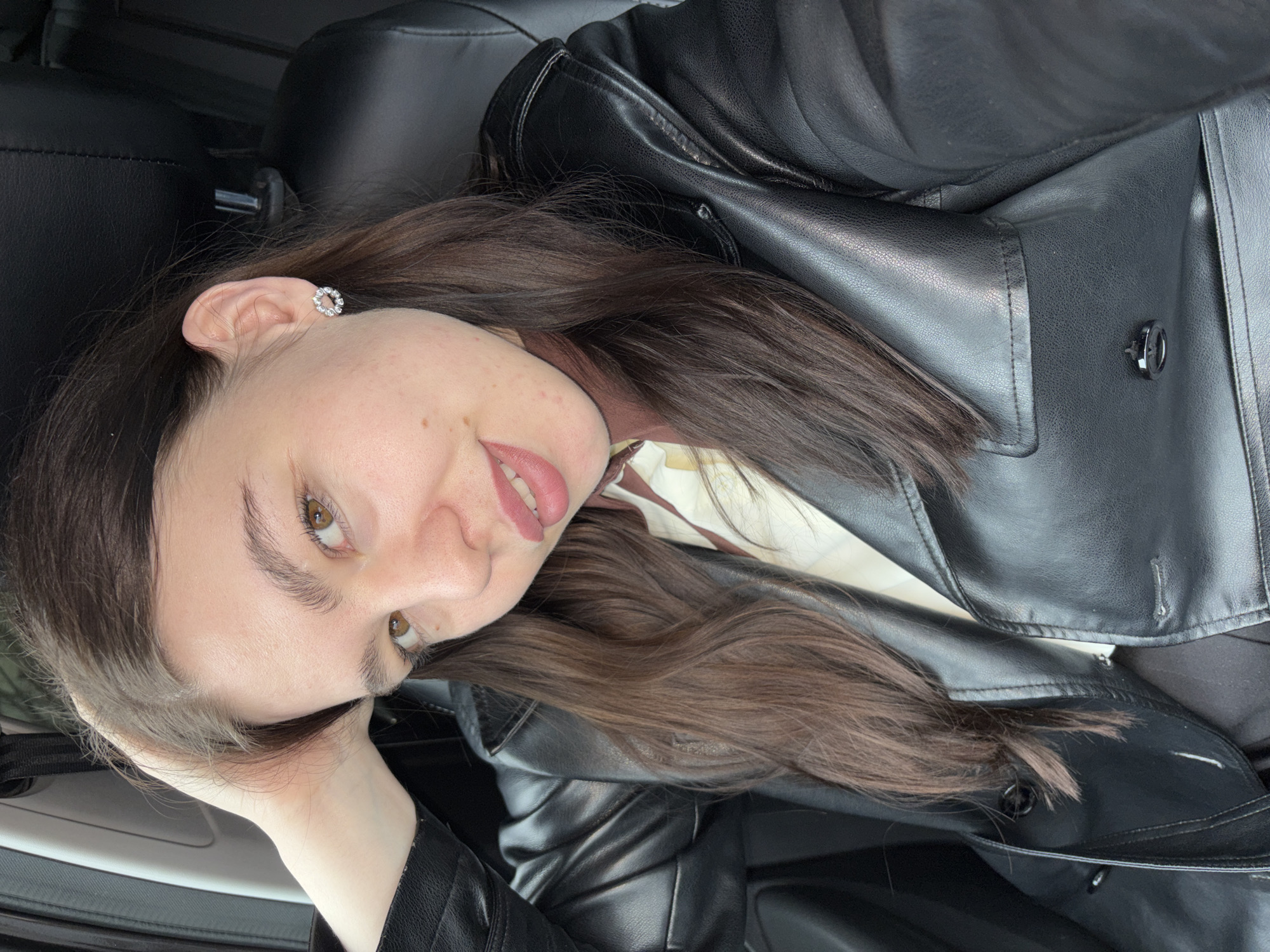 A young woman with long, dark brown hair, fair skin, and light brown eyes is depicted in a close-up, rotated portrait, likely taken inside a car in Perm, Russia. She has visible freckles on her cheeks and nose and is wearing light pink lipstick, with a soft, subtle smile on her face. Her dark eyebrows are well-defined.
She is dressed in a black leather or faux leather jacket, which features prominent stitching and a dark metal button or clasp. A light-colored top with a brown pattern is visible beneath the jacket's collar. On her visible ear, she wears a small, circular stud earring adorned with sparkling clear stones. Her left hand is gently placed near her head, touching her hair.
The background consists of dark, likely leather, car interior elements, including a headrest and seat upholstery, confirming the setting. The lighting is soft and natural, suggesting the photograph was taken during the daytime. No text is visible in the image. The overall impression is one of relaxation and calm.
