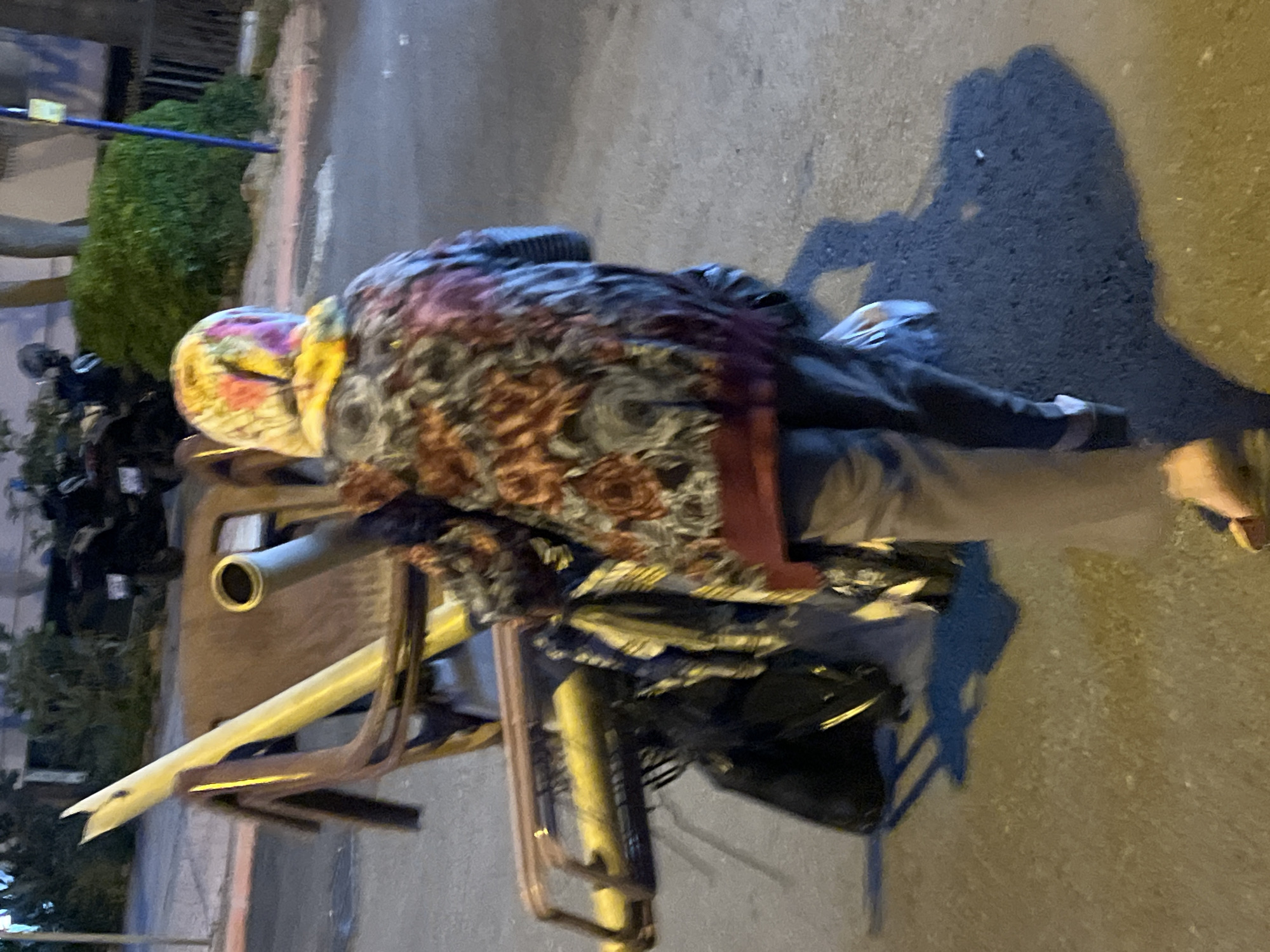 A person wearing a headscarf and floral top is positioned on top of a heavily loaded wheeled platform. The platform appears to be makeshift, constructed from metal tubes, chairs, and an industrial metal pipe, and supports multiple bags. The person is walking on the sidewalk in Alanya, Turkey, as suggested by the surrounding environment. A shadow suggests the person is elderly. Other items are visible on the platform, obscured by the bags and metal frame. There is vegetation visible, and a street with other vehicles parked can be seen in the background.