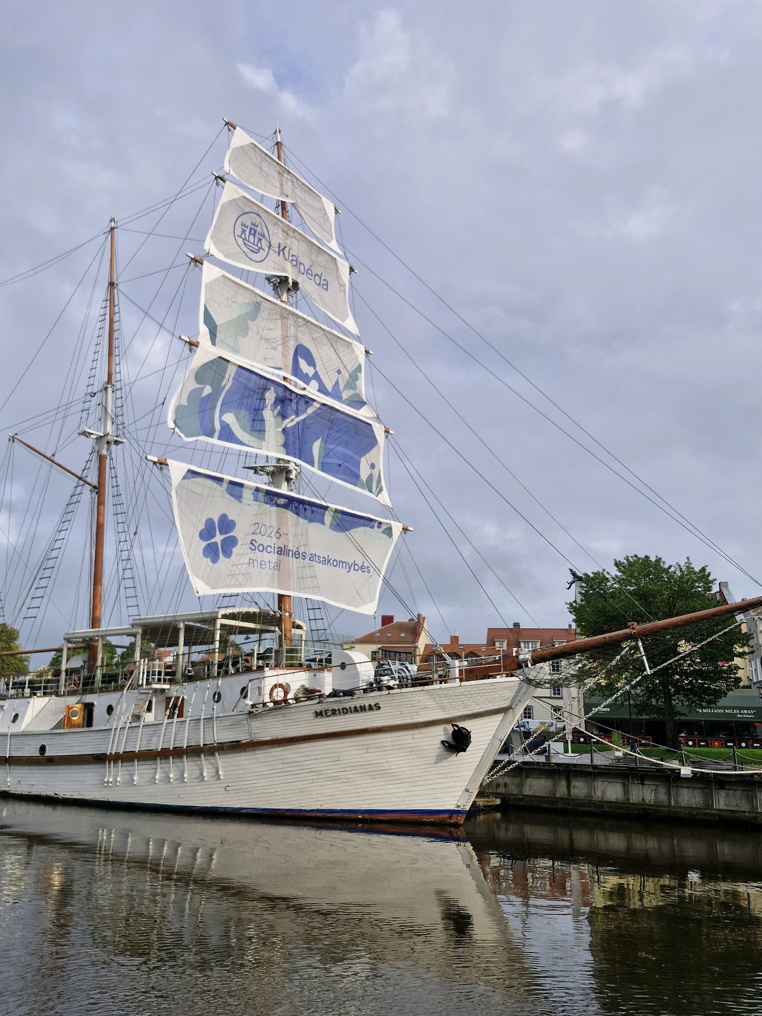 A white sailing ship named "MERIDIANAS" is docked in a harbor, likely in Klaipėda, Lithuania, given the inscription on its sails. The ship has multiple masts and is adorned with large white sails displaying blue floral patterns and text. One sail prominently shows the words "2026 Socialinės atsakomybės metai" which translates to "2026 Year of Social Responsibility," along with a blue four-leaf clover emblem. The ship's hull is white with a blue waterline, and it is reflecting in the calm, dark water of the harbor. In the background, buildings with red-tiled roofs are visible, suggesting an urban waterfront. The sky is overcast with diffused light. The scene appears to be a static, daytime view of the ship at rest.