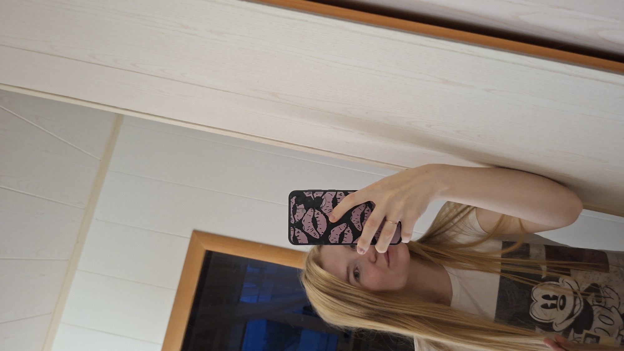 The image shows a young woman with long blonde hair taking a selfie in a mirror. She is wearing a white t-shirt with a Mickey Mouse graphic. She is holding a smartphone with a black case decorated with pink lips. The background consists of paneled walls and a wooden frame, possibly of a window or a doorway. The scene is indoors.