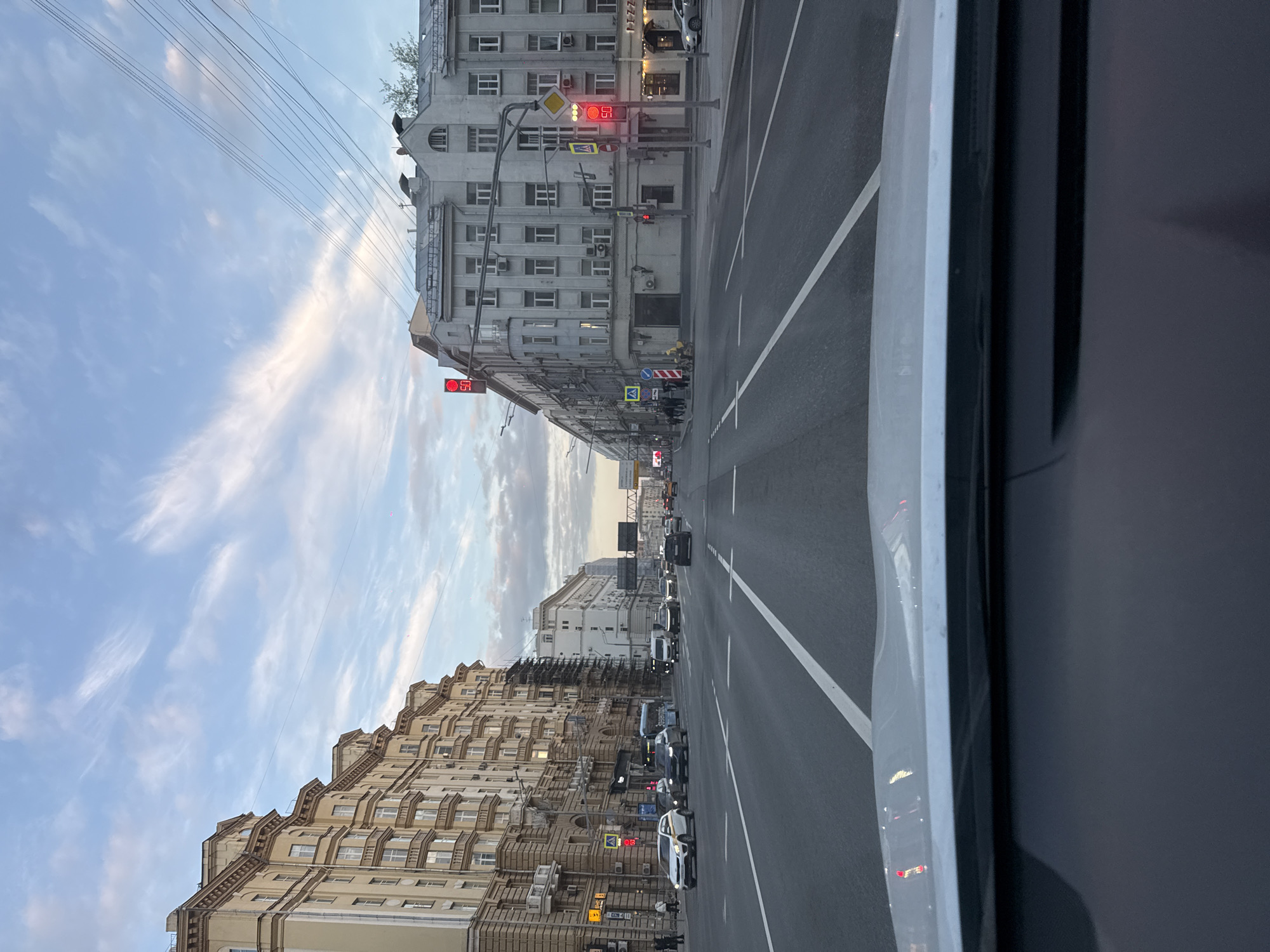 The image shows a street scene in Moscow, Russia. The view is from inside a vehicle, looking down a wide avenue lined with buildings of various architectural styles. The sky is partly cloudy. Traffic is present on the road, including cars. Traffic lights are visible, some showing red. Buildings are mainly light-colored, with multiple windows. Wires are overhead. Road markings include white lines delineating lanes.