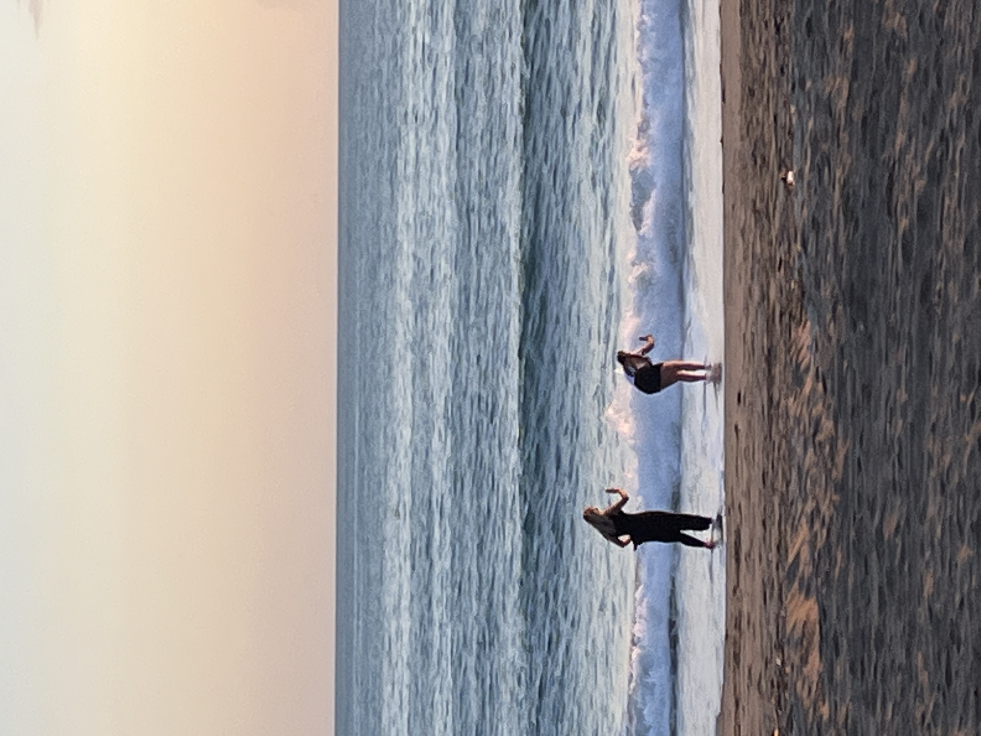 Two people are standing on the beach in Alanya, Turkey. The water line is just at their feet. The tide is coming in. The sky is pale. The sand is dark.