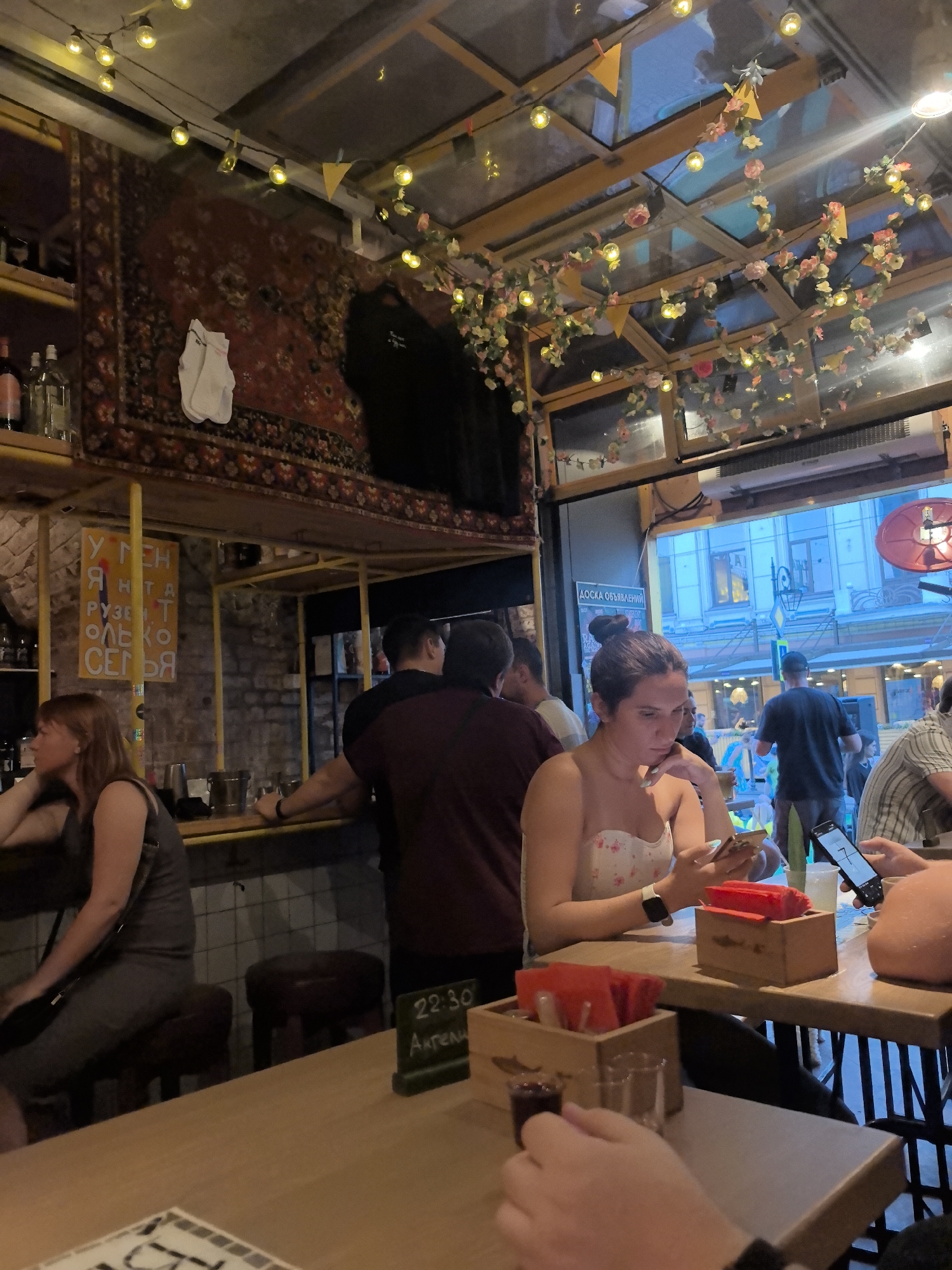 The image shows the interior of a dimly lit bar or cafe, possibly in Nizhny Novgorod, Russia, given the Cyrillic text visible on a sign and a chalkboard. The decor includes a large, ornate rug hanging on the wall, string lights with yellow bulbs, and artificial flowers. Several people are present, with some seated at a bar and others standing. In the foreground, a woman with dark hair pulled into a bun is looking down at her phone. To her right, another person's hands are visible, holding a phone. A table in front of them holds red napkin dispensers and small glasses. In the background, other patrons are visible, and the scene suggests a casual, social atmosphere. The exterior of buildings is visible through an open doorway, with blue lighting and some street activity.