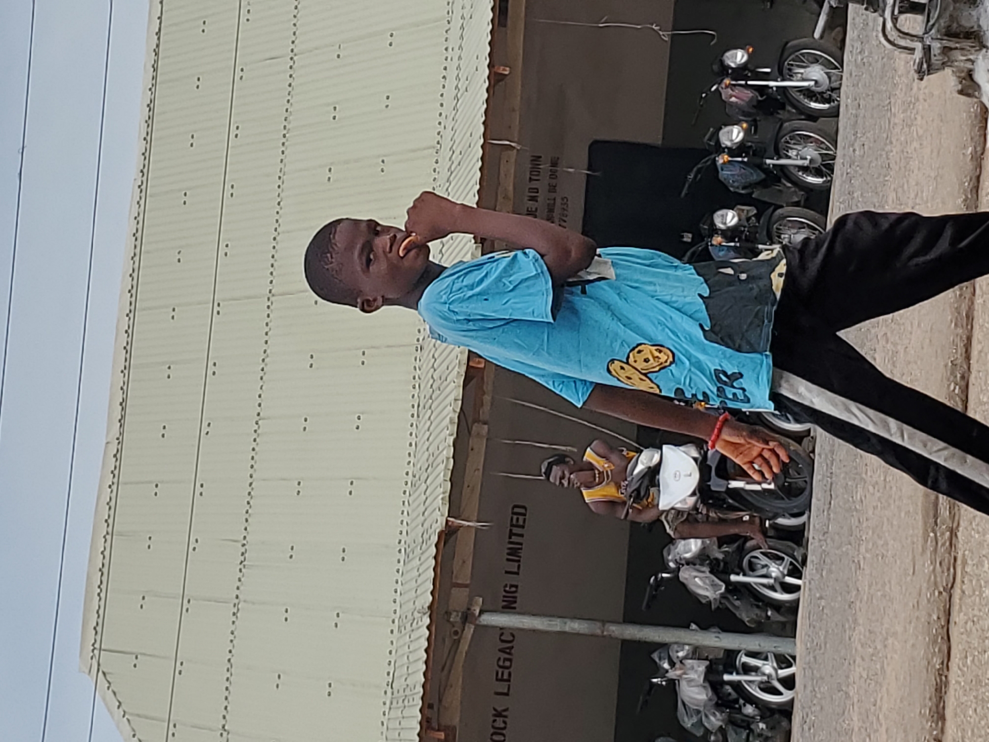A young person in a light blue T-shirt with a cookie pattern and black pants is eating or touching their mouth while standing outside of a building with the inscription "OCK LEGAC NIG LIMITED" and "JE NO TOWN" (likely a business name and location). Multiple motorcycles are parked nearby. Another person is seated on a motorcycle in the background. The location appears to be in a city, based on the presence of power lines and a multi-story building. The city is likely Mararaba, Nigeria, as indicated by the Nigeria (NIG) abbreviation on the building.