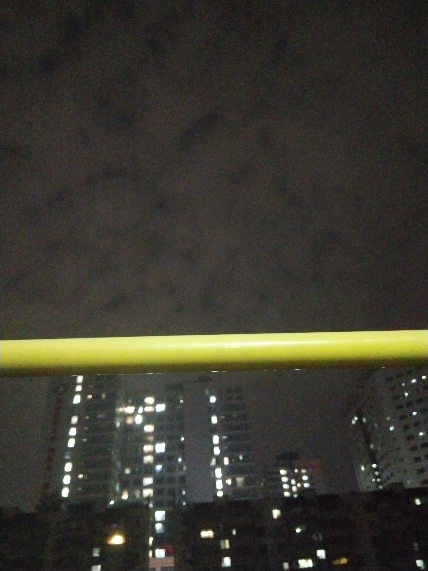 A nighttime shot taken in Yuzhong District, China, shows a blurred view of several tall buildings with illuminated windows, suggesting residential or commercial use. A yellow horizontal bar, likely a railing or part of a structure, is in the foreground. The sky above is dark and possibly cloudy. The buildings' lights create a bokeh effect due to the camera's focus.