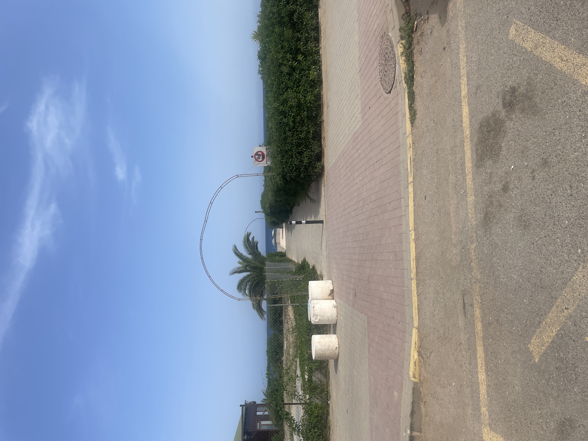 The image shows a pathway leading towards the sea in Lújar, Spain, under a clear blue sky with sparse clouds. The pathway is paved with red and gray bricks, bordered by a yellow curb and a paved road in the foreground. To the left of the pathway, there are three cylindrical concrete bollards and a low green hedge. Further back on the left is a small wooden building with a dark green roof. The pathway is framed by an arched metal structure extending overhead and a tall green hedge on the right. A sign with a red circle and a diagonal line, indicating prohibition, is visible on the right side, mounted on a pole. Beyond the hedge, the sea is visible, with two beach umbrellas in the distance. The environment appears to be coastal, with vegetation and a sense of transition from an urban or developed area to a natural setting.