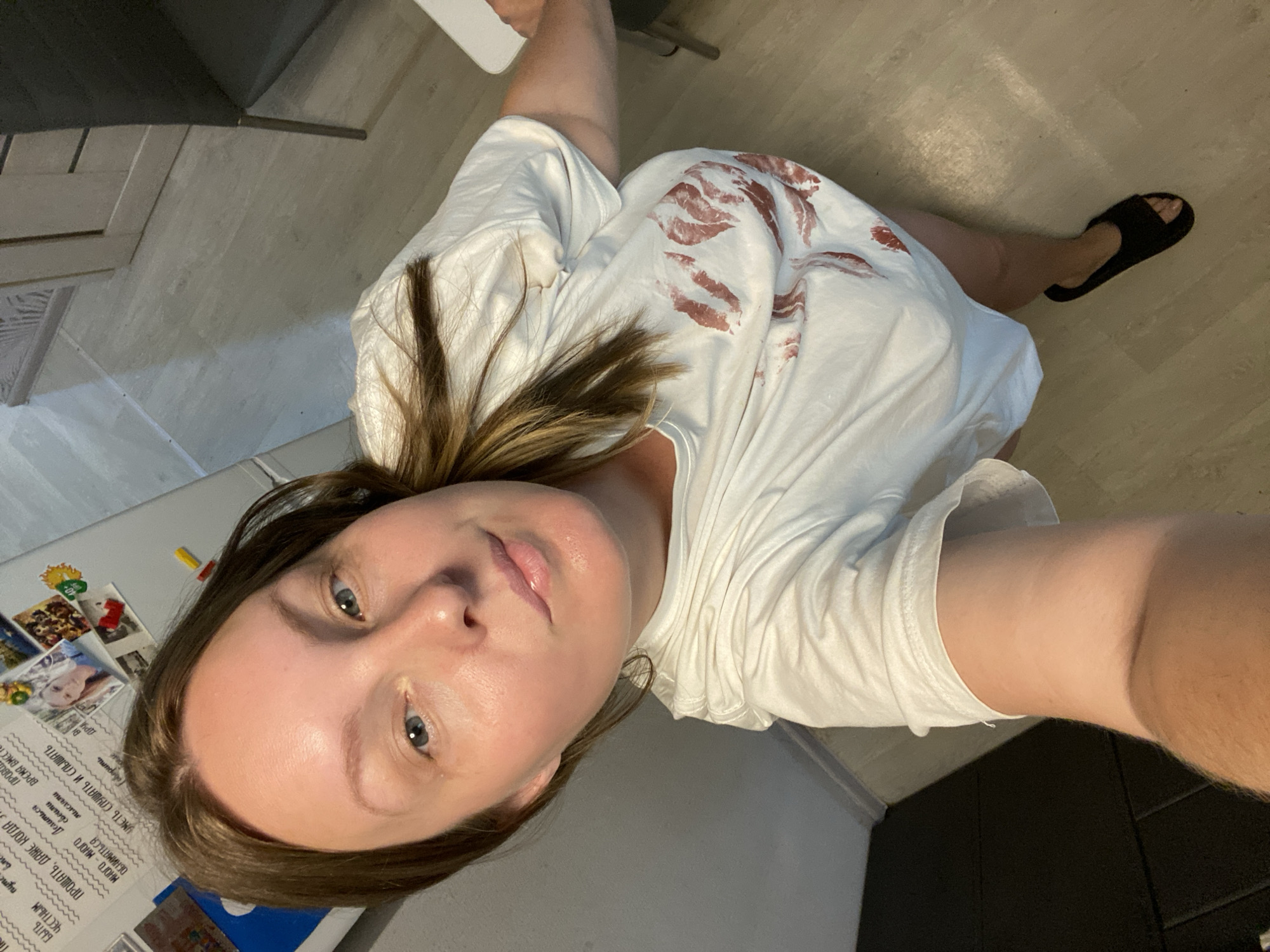 A woman with brown hair, wearing a white t-shirt with red lipstick marks, stands in a room. The floor is a light-colored wood-like laminate. In the background, there is a refrigerator with magnets and photos on it. A black chair is visible to the right of the woman. The woman is looking at the camera. It is not possible to determine the specific location within the city of Noviy, Russia.