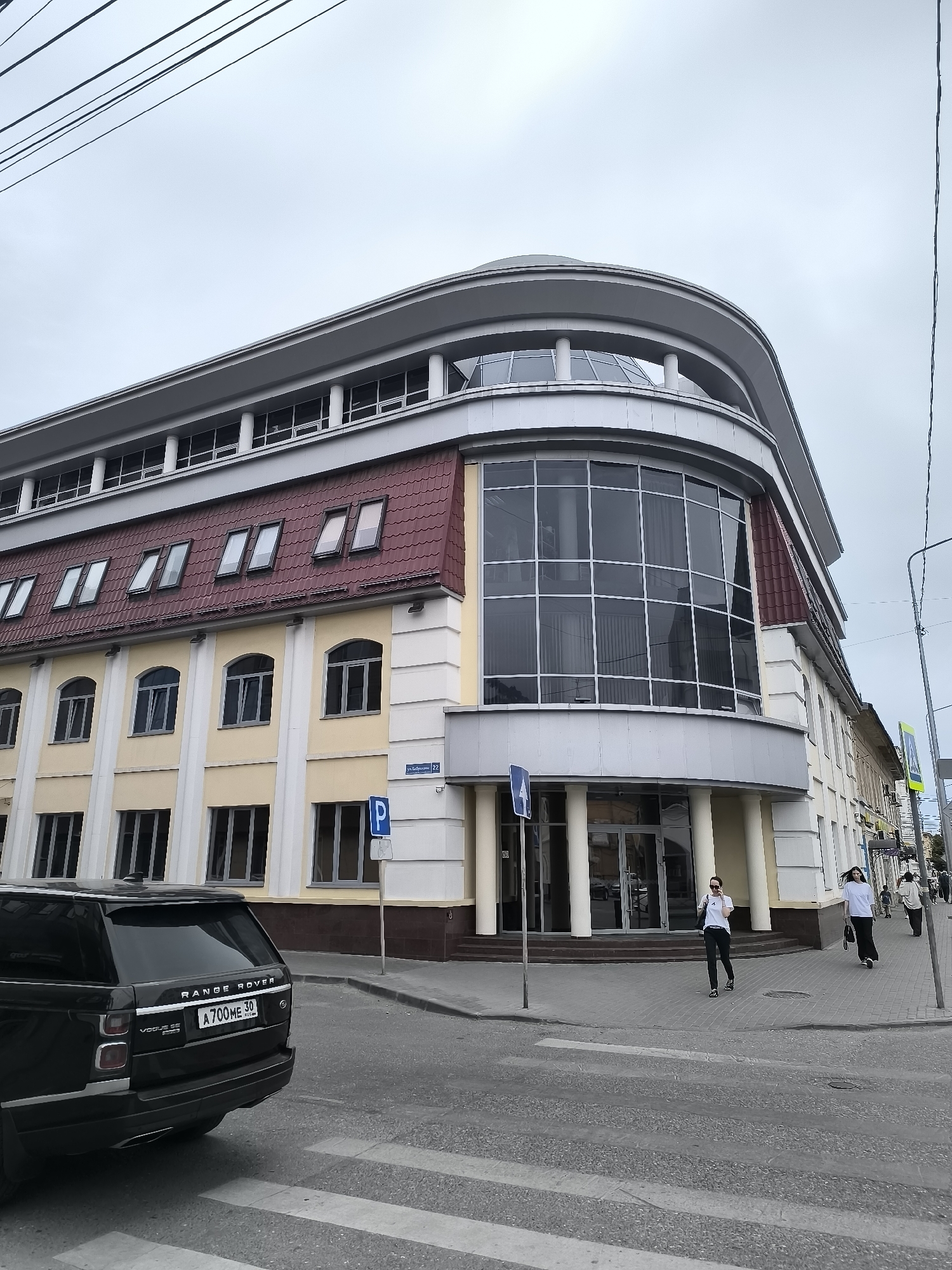 The image captures a street view in Astrakhan, Russia, focusing on a modern, multi-story building with a rounded facade. The building features a red tiled mansard roof with several dormer windows. The main facade is painted a pale yellow with white architectural details and a large, curved glass section on the corner.
In the foreground, a black Range Rover Vogue SE is parked on the left, its license plate reading "A700ME 30 RUS". A pedestrian crossing is visible in front of the vehicle.
On the sidewalk to the right of the building, two women are walking. One woman, wearing a white t-shirt and black pants, is walking towards the camera, while another woman in a white t-shirt and black pants is walking away from the camera. Further down the street, a few more pedestrians are visible, as well as a traffic light and street signs. The sky is overcast, suggesting a cloudy day. A street sign on the building reads "ул. Бабушкина 22".