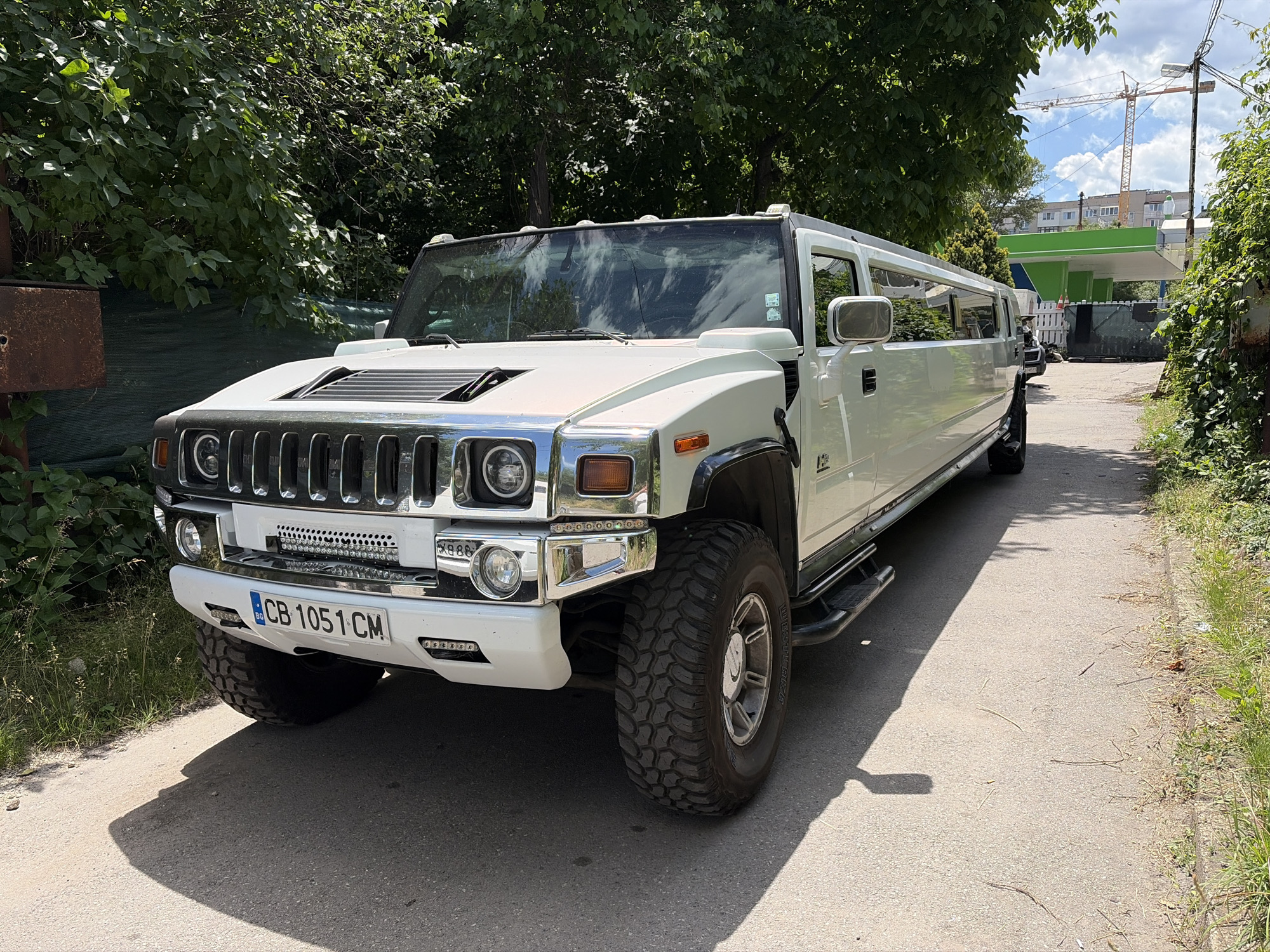 A white, stretched Hummer H2 limousine is parked on an asphalt road in a city. The limousine is facing the viewer, with its front end slightly angled to the left. The vehicle has large, knobby tires and a prominent chrome grille. A license plate reading "CB 1051 CM" is visible on the front bumper. The limousine is flanked by overgrown green foliage and trees on both sides, suggesting a somewhat neglected or naturalized urban environment. In the background, a tall yellow construction crane is visible against a blue sky with scattered clouds. A low-rise building with a bright green roof is also present. The overall impression is of a sunny day in an urban setting, possibly on the outskirts or in an industrial area, given the presence of the construction crane and the overgrown vegetation.