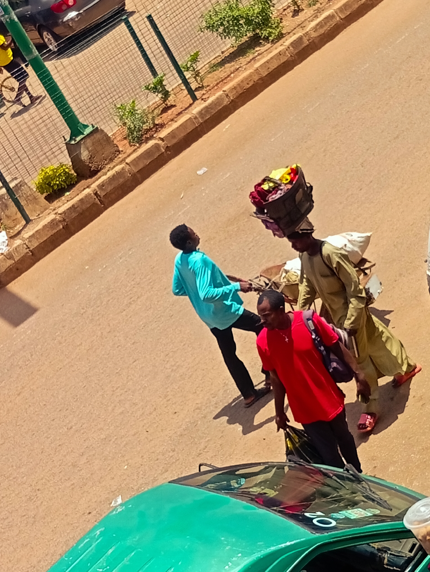 In Bukuru, Nigeria, three individuals are depicted on a paved road. A person in a turquoise shirt walks away from the camera, their head turned to the right. To their left, a person in a red shirt walks towards the camera, carrying a purple bag over their shoulder and holding a black bag in their left hand. To the right, a person wearing a light green tunic and trousers carries a large basket filled with colorful items on their head, with a white sack strapped to their back and a wooden cart pulled behind them. The environment is a street scene with a sidewalk and a metal fence bordering a patch of vegetation. A green vehicle is partially visible in the foreground, with its windshield and part of the hood in view.