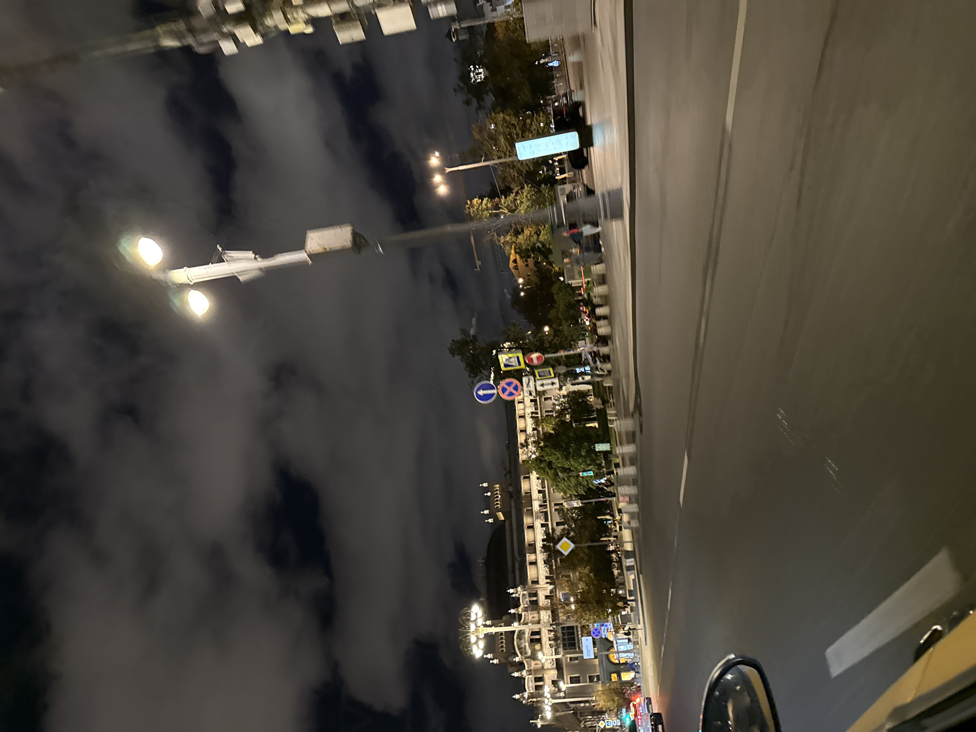 The image captures a nighttime view of a city street in Moscow, Russia, likely from a moving vehicle. A grand, illuminated building with ornate architecture stands prominently on the left side of the frame. Streetlights illuminate the cloudy sky and the roadway. Various traffic signs, including directional arrows and a yield sign, are visible along the sidewalk. Several trees line the street, their foliage illuminated by the artificial lights. The road itself shows tire marks, indicating traffic. In the foreground, the side mirror of a vehicle and a portion of its yellow body are visible. The overall atmosphere is urban and somewhat moody due to the nighttime setting and cloudy skies.