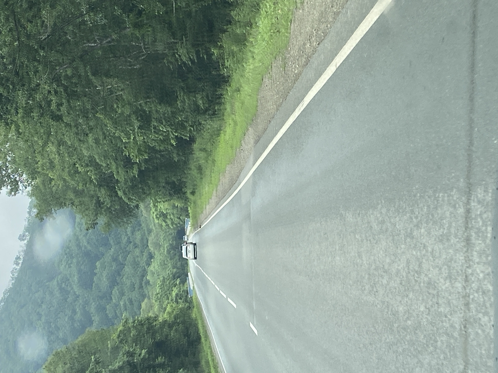 The image depicts a two-lane asphalt road in a rural, mountainous area, likely Ryazanovka, Russia. The road is bordered by dense, green foliage on both sides, with a large, tree-covered mountain range in the background. The sky is overcast, suggesting a cloudy or possibly rainy day, as evidenced by faint circular water spots on the camera lens.
There are two white cars visible on the road, driving in the same direction as the camera. The lead car appears to be a sedan, followed by another vehicle behind it. The road has white lane markings, indicating a divided highway or a road with clear directional guidance. The presence of guardrails and possibly a small blue bridge or structure in the distance suggests infrastructure common to such roadways. The ground beside the road is covered in grass and gravel. The overall scene conveys a sense of travel through a natural, verdant landscape.