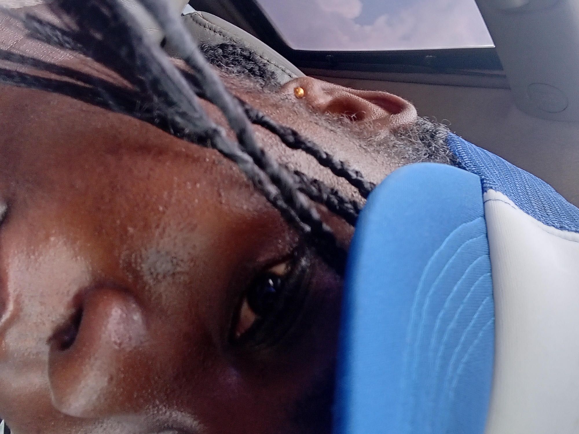 This is a close-up, eye-level, indoor photograph of a person's face, partially obscured by a blue and white baseball cap. The person appears to be of African descent, with braided hair visible on the left side of the frame. A small, gold stud earring is visible in the left earlobe. The lighting suggests the photo was taken during the day, possibly in a vehicle, given the reflection of the sky in the background. The image is taken in Adoma, Nigeria.