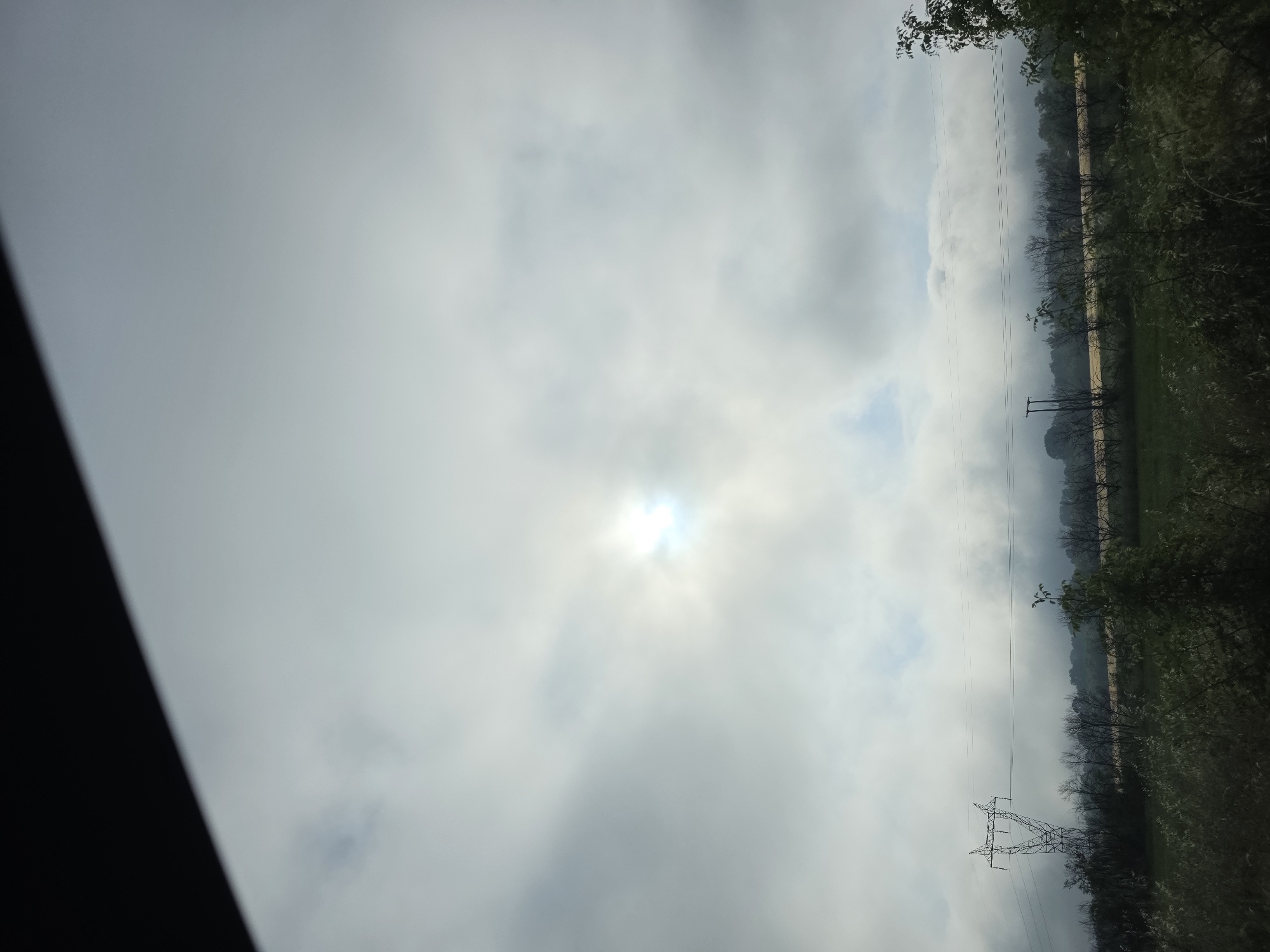 The image captures a rural landscape under a cloudy sky, likely taken from inside a vehicle due to the dark, diagonal element in the top left corner. The sky is predominantly covered by a layer of light grey clouds, with the sun brightly breaking through a less dense area in the upper center, creating a strong glare.
Stretching across the middle ground are multiple horizontal power lines. A large metal electricity pylon (lattice tower) is visible on the lower left side, supporting some of these lines. Further to the right, a simpler utility pole can be seen. Beyond the power lines, a line of trees or dense vegetation extends across the horizon, appearing somewhat muted or silhouetted against the hazy sky. Some trees, particularly in the lower right, show denser foliage, while others in the distance appear more sparse or bare. The ground below the trees seems to be fields or open terrain.
The time of day appears to be midday or early afternoon, indicated by the high position of the sun. The weather is overcast but with moments of sunlight. No specific activity is taking place, and no people are visible. The scene depicts a quiet, expansive view of the countryside. There is no visible text in the image. Given the provided context, the location is Dolna Mahala, Bulgaria.