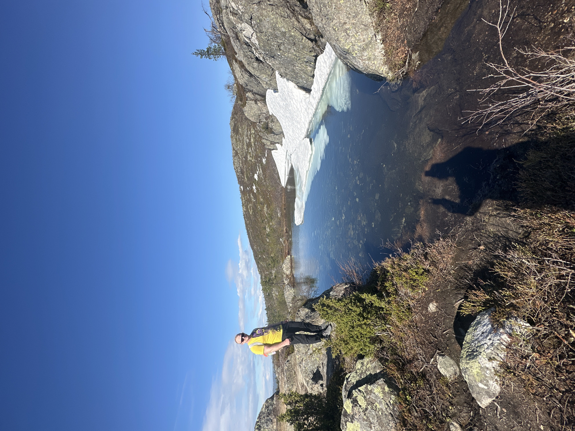 A man wearing a yellow shirt and black pants stands on a rocky ledge overlooking a small body of water. Patches of ice float in the water near a rock face. The sky is clear blue. The location is likely near Kandalaksha, Russia, given the landscape and presence of glacial features like the ice patch. The man is facing the camera and appears to be posing. The environment consists of rocks, low-lying vegetation, and a small lake or pond. The sun is shining brightly, casting a shadow of the man onto the ground near the water's edge.