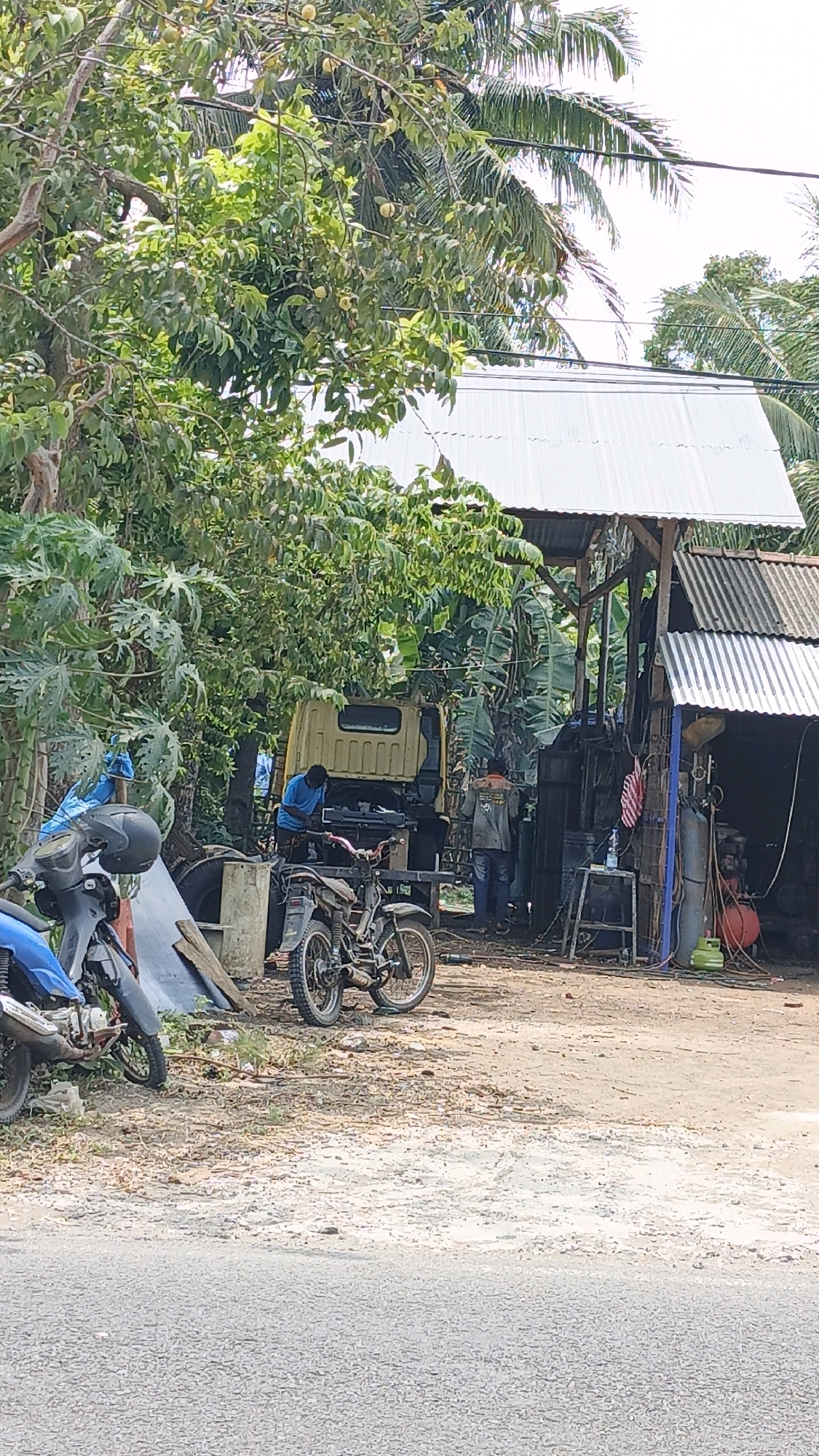 In Desa Wonoharjo, Indonesia, a modest outdoor workshop operates under a corrugated metal roof supported by wooden posts. A yellow truck is positioned near the workshop, with a person in a blue shirt appearing to work on its engine. Another individual, wearing a jacket and jeans, stands nearby. Two motorcycles are parked in front of the workshop, one blue and one a dirt bike. The ground is unpaved and appears dusty. Trees and vegetation surround the area, suggesting a rural or semi-rural setting. The sky is overcast.