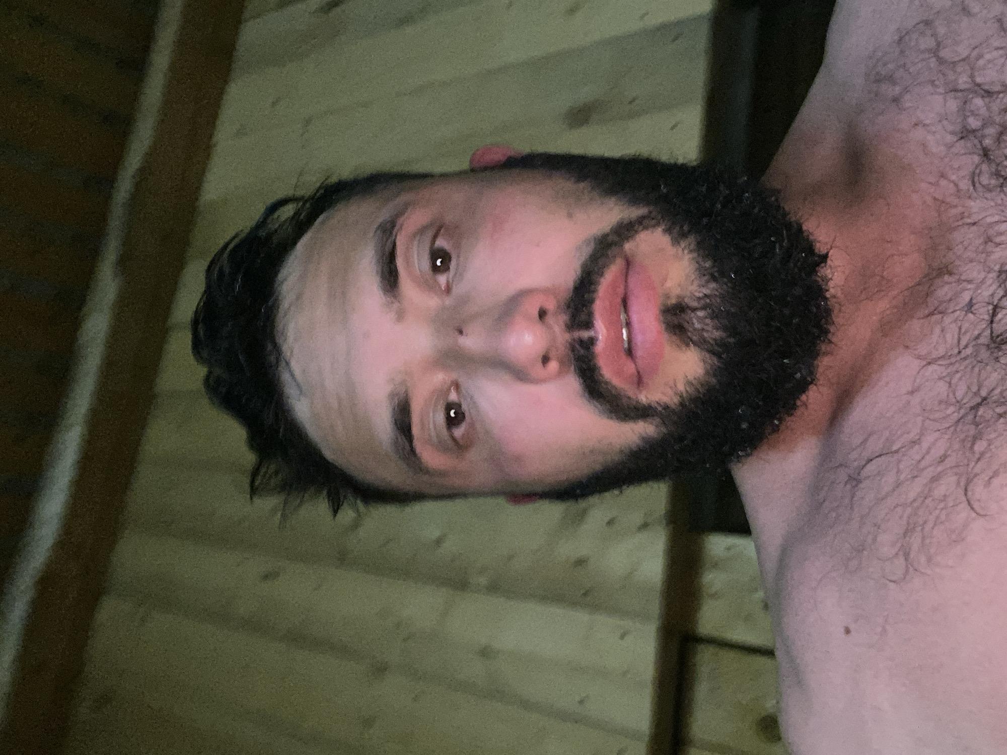A close-up, chest-up selfie of a man with a beard and dark hair is taken from a low angle, inside a wooden sauna. The man is looking directly at the camera, his expression neutral. The wooden walls of the sauna are visible in the background, with vertical planks of wood. The lighting is dim and warm, possibly from the sauna's heat or artificial lighting. The man appears to be shirtless, with visible chest hair. His skin has a flushed tone, consistent with being in a hot environment like a sauna.