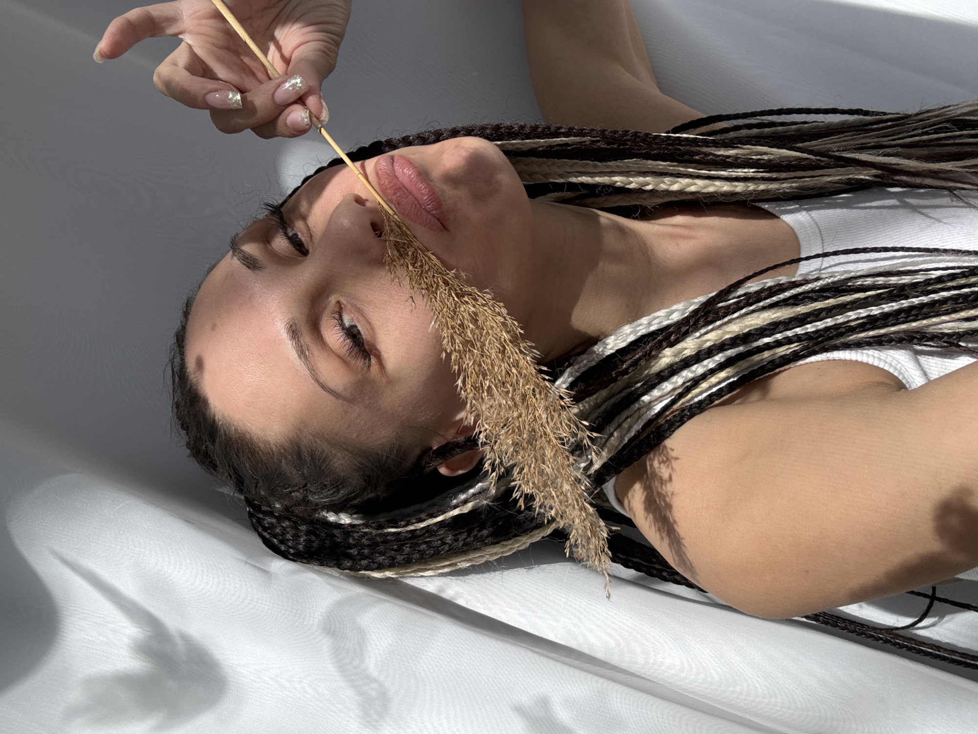 An artistic, high-angle close-up portrait shows a person lying on their back on a white fabric surface, illuminated by strong natural light that casts distinct shadows. The setting is indoors, likely in Rusanovo, Russia, though specific location cues are not visible.
The primary subject is a female-presenting individual with dark brown, light brown, and white/blonde synthetic braids or dreadlocks fanned out around her head and shoulders. She is wearing a simple white ribbed tank top. Her head is slightly tilted, and she looks upwards with a contemplative or serene expression. Her lips appear to have a soft, natural-toned gloss.
One of her hands is visible in the upper left of the frame, holding a thin, light-colored stick with a plume of dried, feathery grass or reeds. Her nails are manicured with a light, possibly iridescent or glittery polish. The dried plant extends downwards, resting near her nose and mouth, partially obscuring her right cheek.
The white fabric surface she lies on is subtly textured with folds, and the harsh, directional lighting creates sharp shadows across it, suggesting bright sunlight from an unseen window or opening. These shadows imply a specific time of day, likely morning or late afternoon, when the sun is at a lower angle. No text is visible in the image. The overall impression is one of natural beauty and an artistic study of light and form.