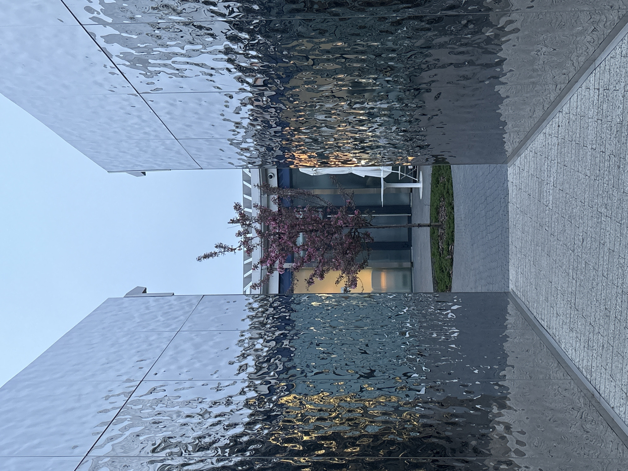 The image shows a narrow passageway between two buildings. The walls of both buildings are covered in reflective, textured metal panels. In the center, between the buildings, is a flowering tree. In the background, there is a white table and chair set visible in front of a building with large windows. The ground is paved with gray stone. The sky is a pale blue. The location is Высокая Гора, Russia.
