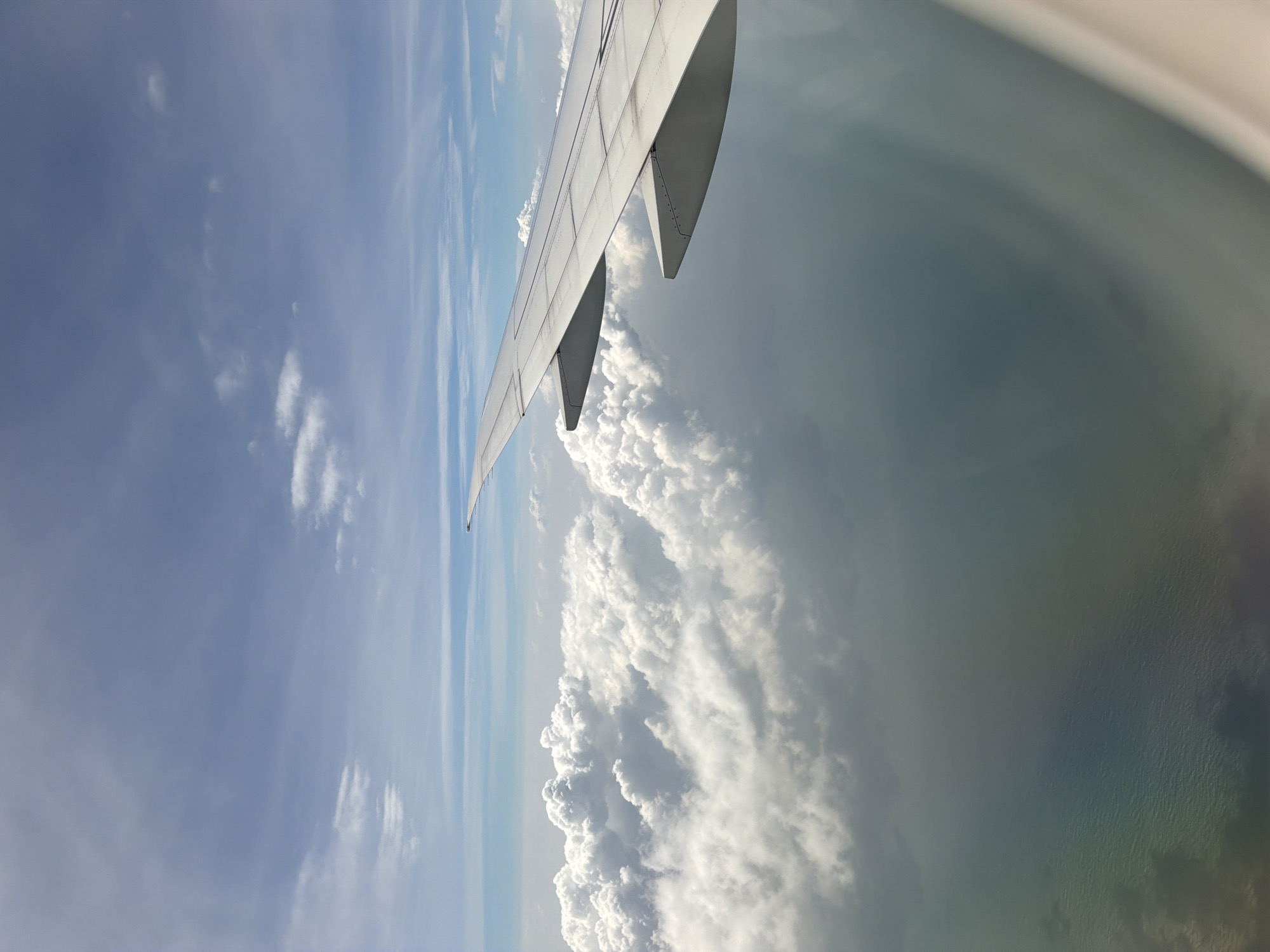 The image is an aerial view from an airplane window, showing a cloudscape above the ocean. The right side of the image is dominated by the wing of the airplane, which is white and appears to be a modern jetliner. Below the wing and extending to the left are large, fluffy white clouds. The ocean is visible beneath the clouds, appearing as a muted green and blue expanse with some texture suggesting waves. There is a faint prismatic effect visible on the water's surface, possibly a rainbow or light refraction. The sky above is a clear blue with some wispy white clouds. There are no people or specific landmarks visible that would indicate a location within Singapore.