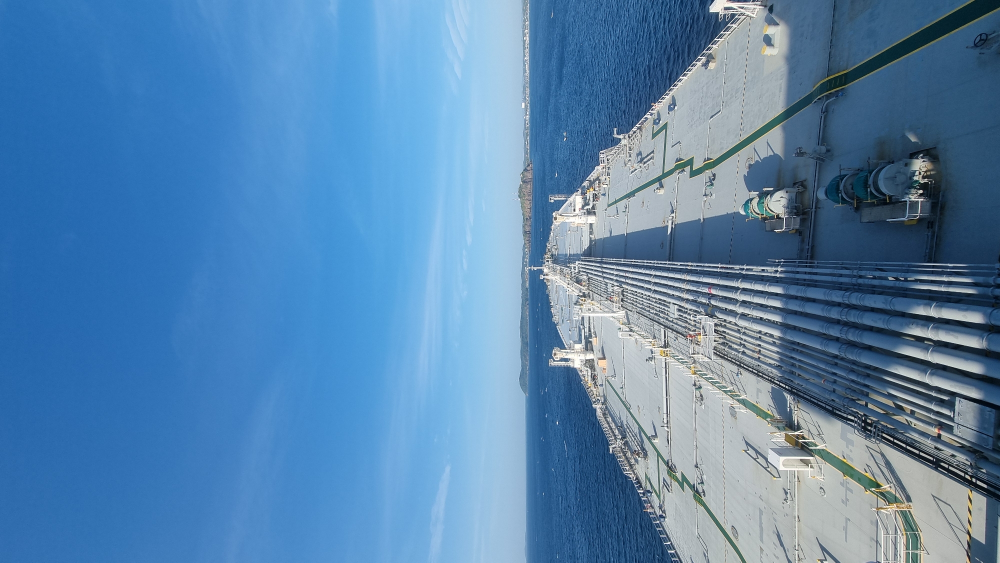 The image shows a wide-angle view from the stern of a large oil tanker sailing on a calm, deep blue ocean. The deck of the tanker is visible, with numerous large pipes running along its length. Several large green winch-like devices are also present on the deck. In the distance, a hazy coastline with hills and some structures can be seen under a clear blue sky with a few wispy clouds. The image depicts a maritime scene, likely in open water. The reference to Trincomalee, Sri Lanka, is not visually supported by the image content, as no specific landmarks or identifying features of that location are present.