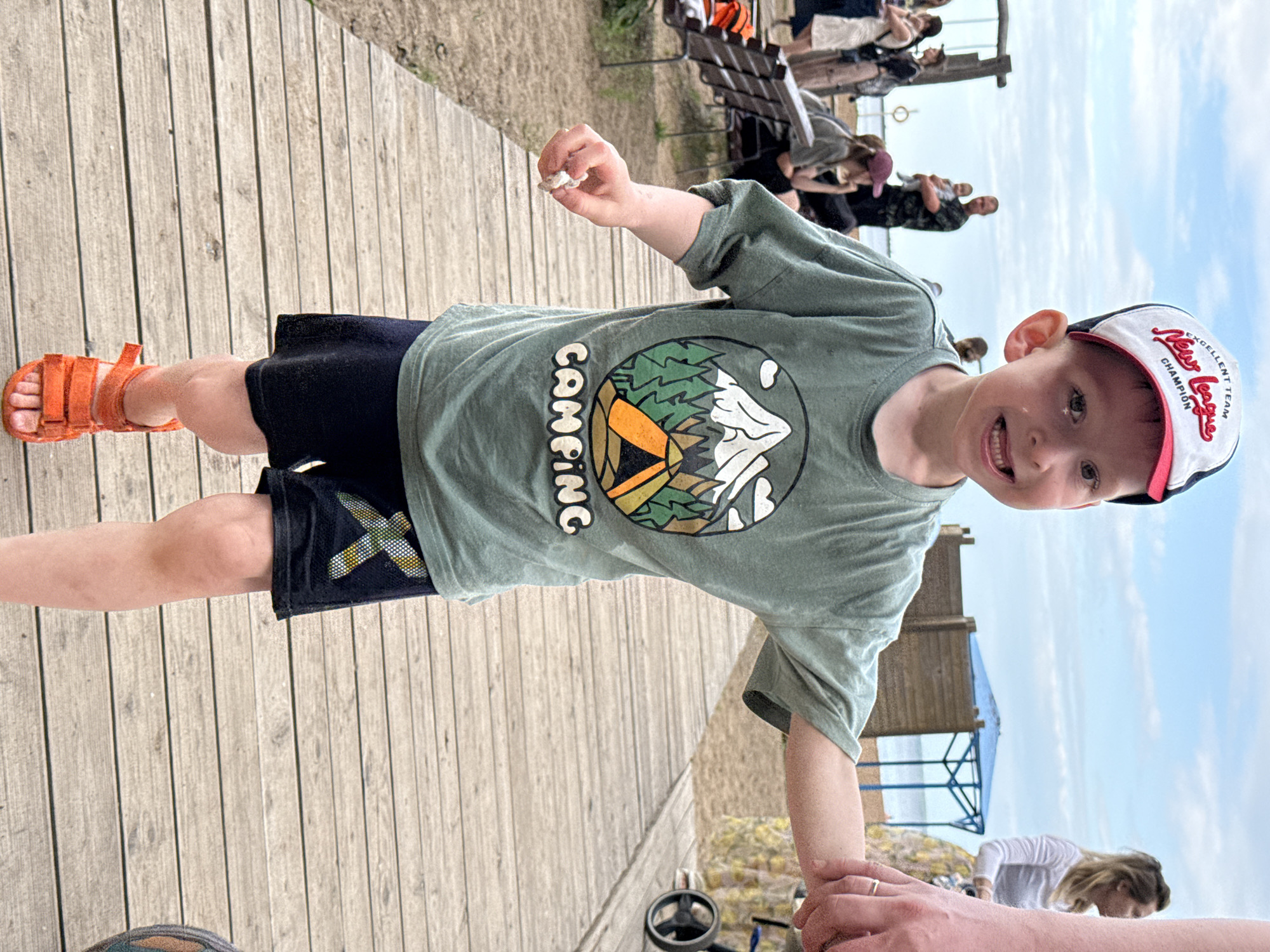 A young boy, wearing a green t-shirt with a graphic of mountains and the word "Camping" on it, and black shorts, is smiling at the camera. He is wearing an orange sandal on his left foot and a baseball cap that says "Excellent Team" and "Champion" on it. He is holding a small, white object in his right hand.
The boy is standing on a wooden boardwalk. In the background, there are other people, some of whom are sitting on benches. There is also a wooden structure and a body of water, likely a lake or the sea. The sky is cloudy. The scene appears to be outdoors, possibly at a beach or a park. The boy's expression suggests happiness and excitement.
The time of day appears to be daytime, given the lighting. The weather seems to be overcast. The location cues suggest a recreational area by the water.