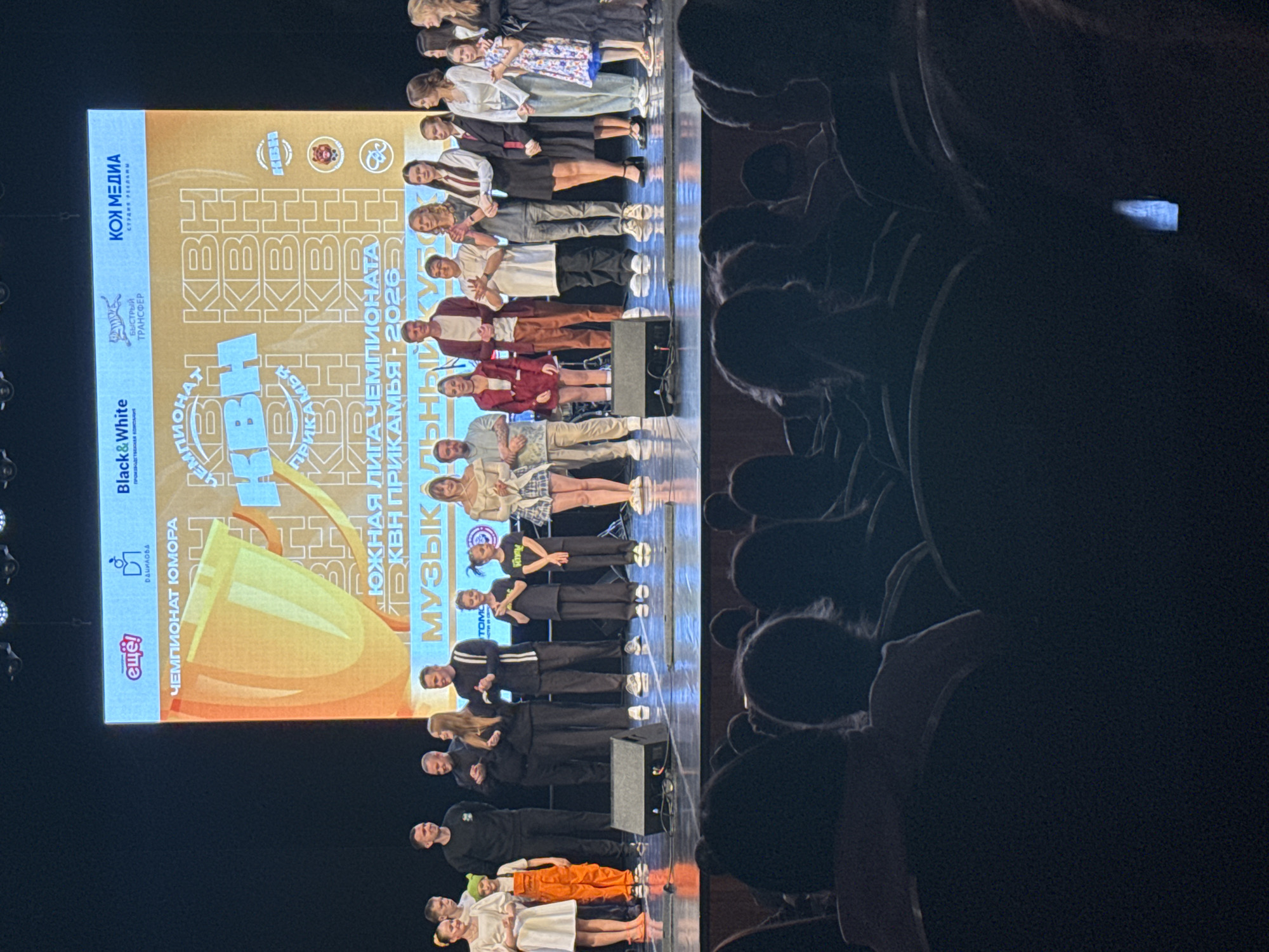 This is a low-angle photograph of a stage performance likely taken from the audience perspective. On the left, a large screen displays logos, including "Black&White," "KOH MEDIA," and "Championship of Humor," with a gold trophy image. Text in Russian indicates the "Southern League Championship" and the years "2026."
A group of people stands on the stage, seemingly arranged in a line or several rows. These appear to be performers or contestants of some kind. The group consists of at least twenty individuals, ranging from children to adults, in various outfits.
The right side of the image shows the backs of audience members' heads, suggesting a crowded venue. The lighting in the venue appears dim, contrasting with the brightly lit stage. The location is likely in the city of Chaikovsky, Russia, based on the Russian text on the screen. The interaction between the people on the stage and the audience is implicit, with the stage set for a performance or competition.