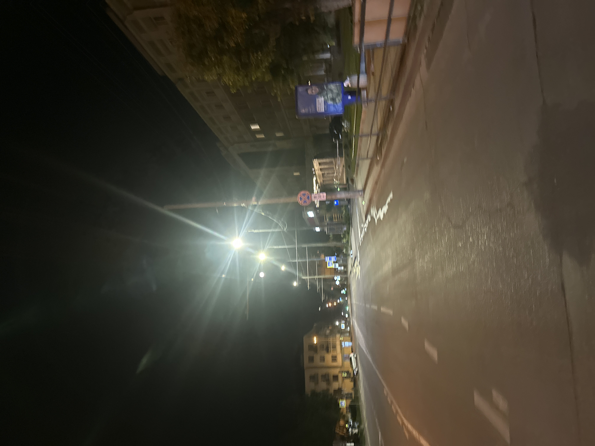The image captures a nighttime street scene in Ternopil, Ukraine, likely on a road with multiple lanes. Streetlights illuminate the asphalt, creating reflections and glare, particularly from multiple light poles visible along the side of the road. On the right side of the road, a multi-story building with illuminated windows is present, and a large advertisement billboard featuring a person in military uniform is displayed. A metal fence runs along the sidewalk in front of the building. On the left side of the road, another multi-story building is visible, appearing lighter in color and with illuminated windows. Several cars are parked or driving on the street. Traffic signs and road markings, including white dashed lines, are discernible. The sky is dark, indicating nighttime, with some visible wires overhead. The overall impression is a typical urban street at night.