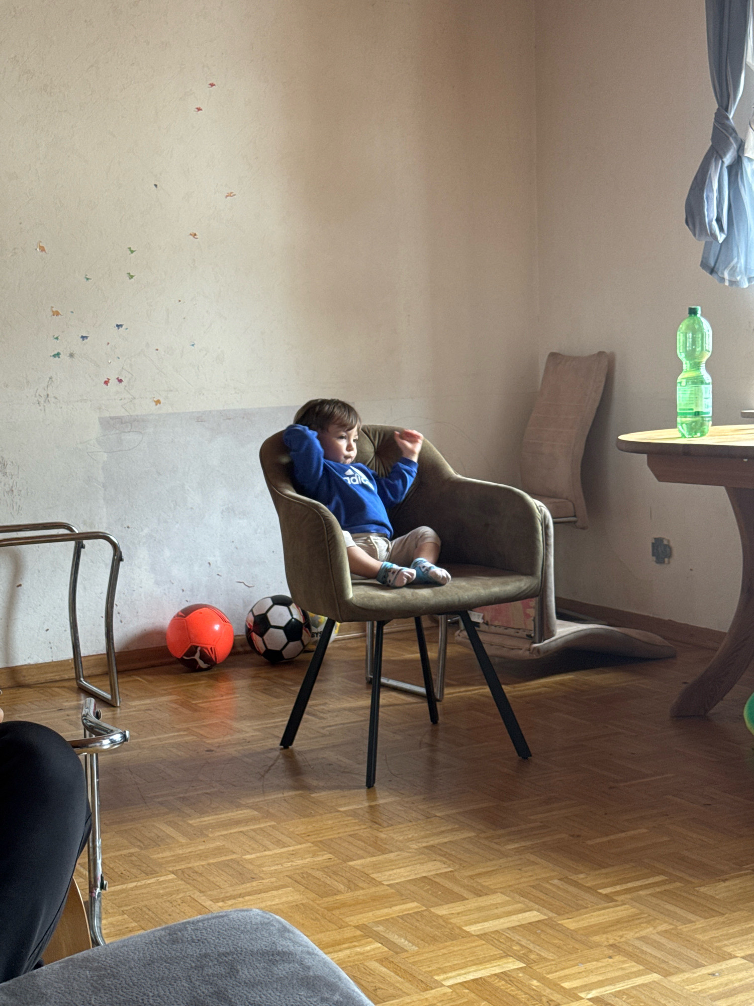 The image depicts an indoor scene, likely a living room in Überherrn, Germany. A young child, appearing to be a toddler, is seated in an upholstered armchair. The child is wearing a blue hooded sweatshirt and light-colored shorts, with socks on their feet. They are looking to the right with their hands behind their head, seemingly relaxed or contemplative.
In the foreground, to the left, is a metallic rack with a curved top. On the floor in front of the armchair are two balls: one orange and one black and white. The floor is made of polished wood parquet. The wall behind the child is textured and appears to have some faint, colorful decals, possibly of small animals. To the right of the armchair, part of a round wooden table is visible, on which rests a green plastic bottle of liquid. Further to the right, a portion of a wall-mounted, beige chair is visible. The overall lighting suggests it is daytime, with natural light entering from the right side of the frame, casting shadows on the floor.