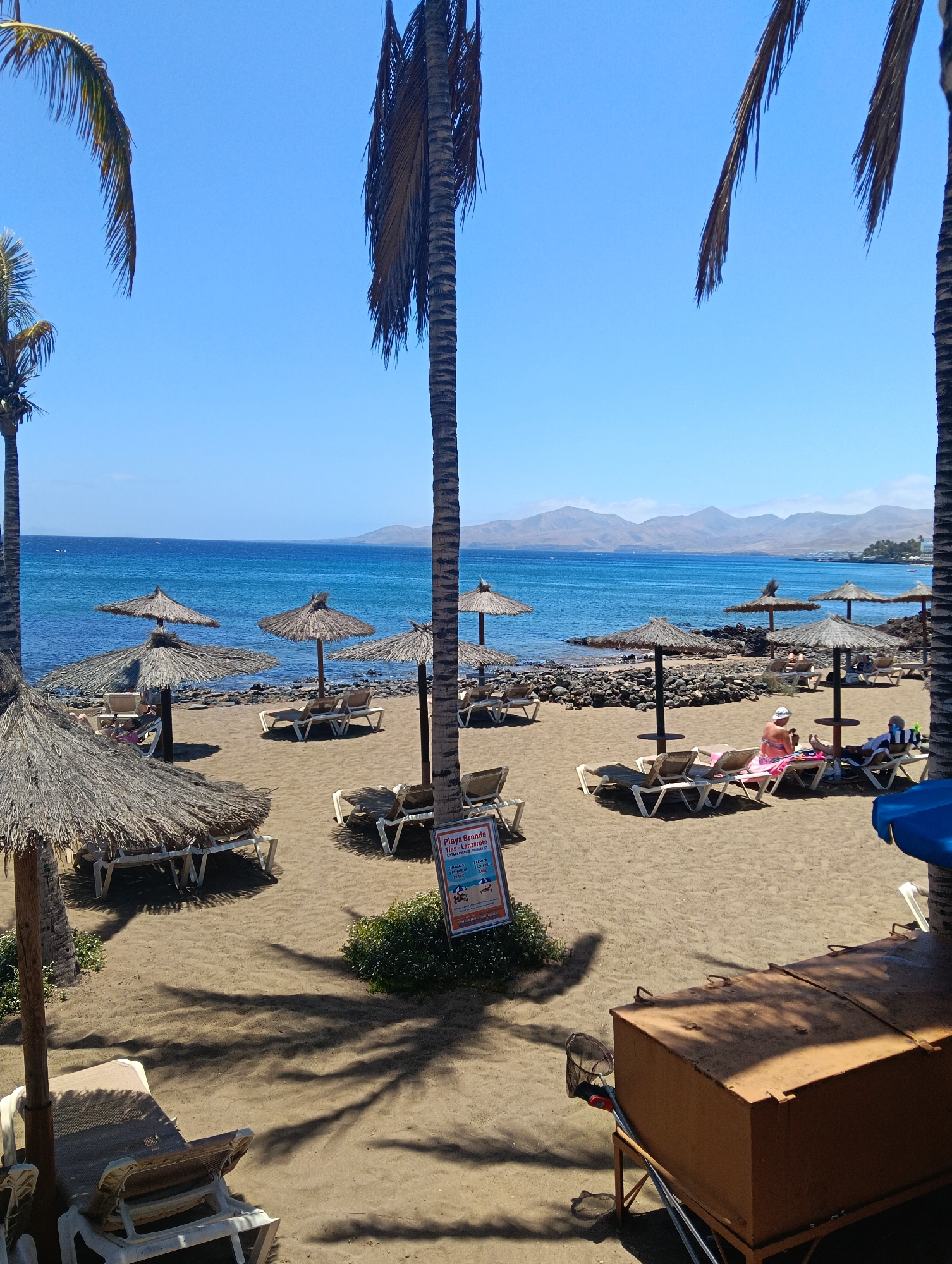 This vibrant image depicts a sunny beach scene at Playa Grande in Puerto del Carmen, Lanzarote, Spain. The sky is a clear, brilliant blue, meeting the calm, bright blue waters of the ocean. In the distance, arid mountains or hills are visible across the water.
The sandy beach is furnished with numerous straw parasols and white beach loungers, many of which are unoccupied. A few individuals are present, enjoying the warm weather. One person wearing a pink top and a white hat is clearly visible relaxing on a lounger under a parasol, facing away from the camera. Other people are further down the beach.
Tall palm trees frame the scene, with their fronds partially visible at the top and left. They cast long, distinct shadows across the sand, suggesting it is either late morning or early afternoon. In the bottom right foreground, a large, rusty-orange metal box on a stand is prominent, with a net leaning against it.
A white sign is visible near the center of the image, attached to a post or palm tree. The text on the sign clearly identifies the location as "PLAYA GRANDE TÍAS - LANZAROTE." Below this, pricing information is displayed, including "1 HAMACA," "2 HAMACAS," and "1 SOMBRILLA," with a price of "7.00 €" visible for some rentals, indicating costs for sunbeds and parasols. The overall mood is one of peace and leisure on a bright, clear day.