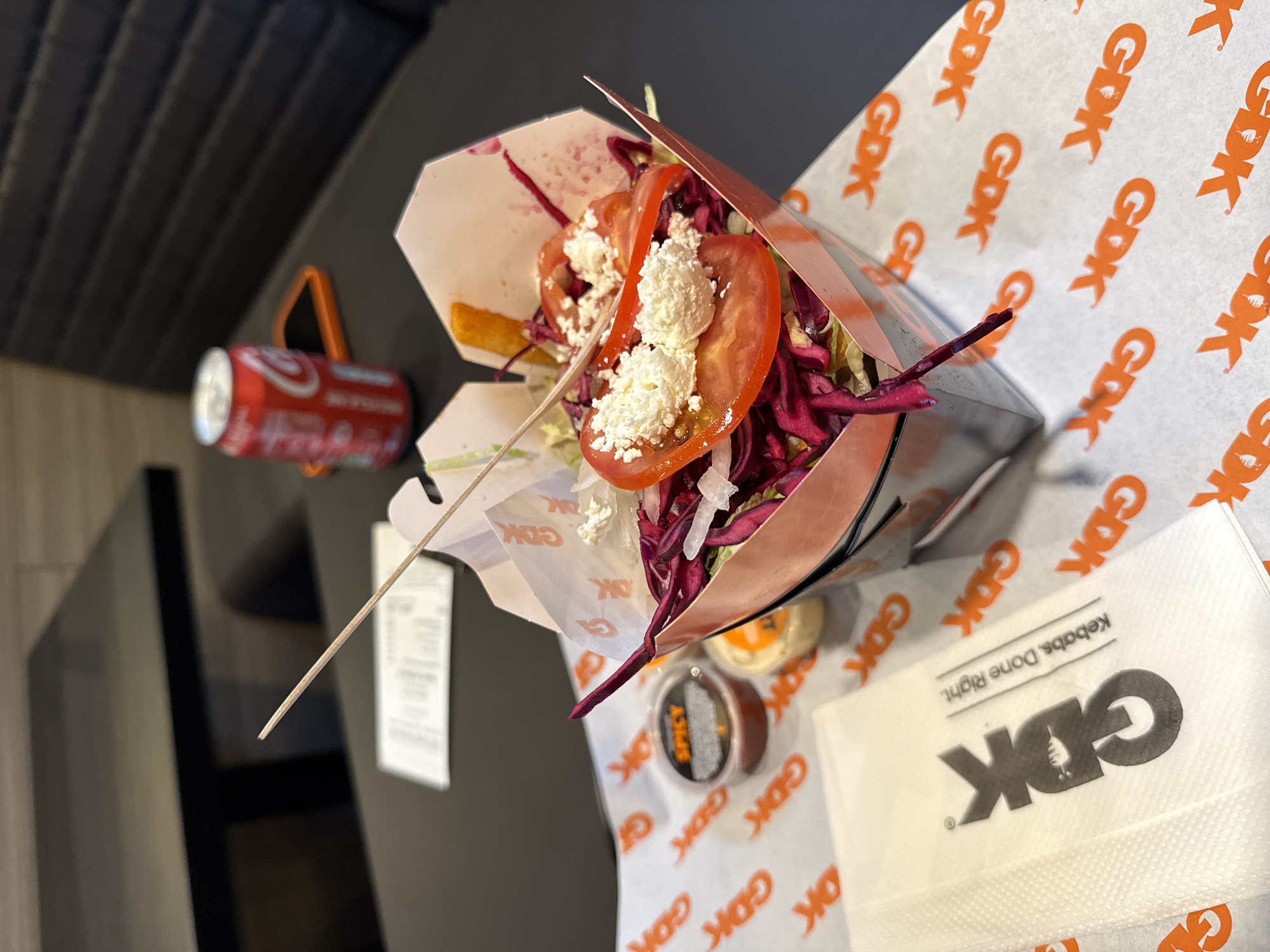 The image captures a German Doner Kebab (GDK) meal. A partly consumed kebab, featuring visible tomato slices, red cabbage, and cheese, sits within a GDK branded container. Accompanying the kebab are small tubs, possibly containing spicy sauce and another condiment. The meal rests on GDK branded paper and a napkin. A can of Coca-Cola is visible in the background.