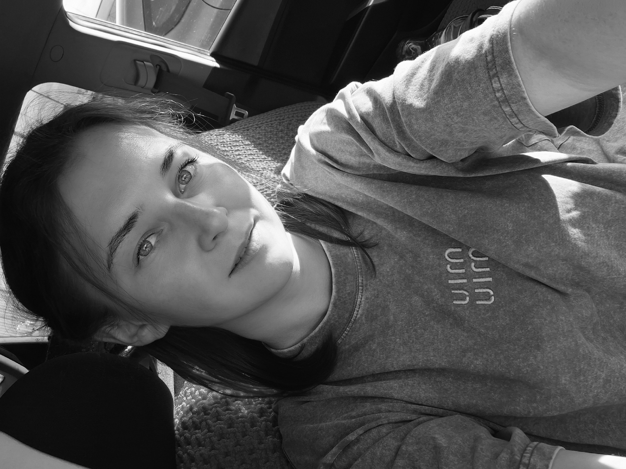A black and white, high-angle, selfie-style photograph shows a woman with light-colored eyes and hair pulled back from her face, sitting in the passenger seat of a vehicle. The woman is wearing a grey t-shirt with a textured weave and a logo on the left breast. Sunlight streams in from the right, illuminating her face and casting shadows. The interior of the car is visible, including the headrest, seatbelt, and door panel. The background is blurred, suggesting a motion shot or shallow depth of field. The image's composition and lighting suggest it was taken during the daytime. The exact location within Kamyennoye, Russia, is not discernible from the visual information provided in the image.