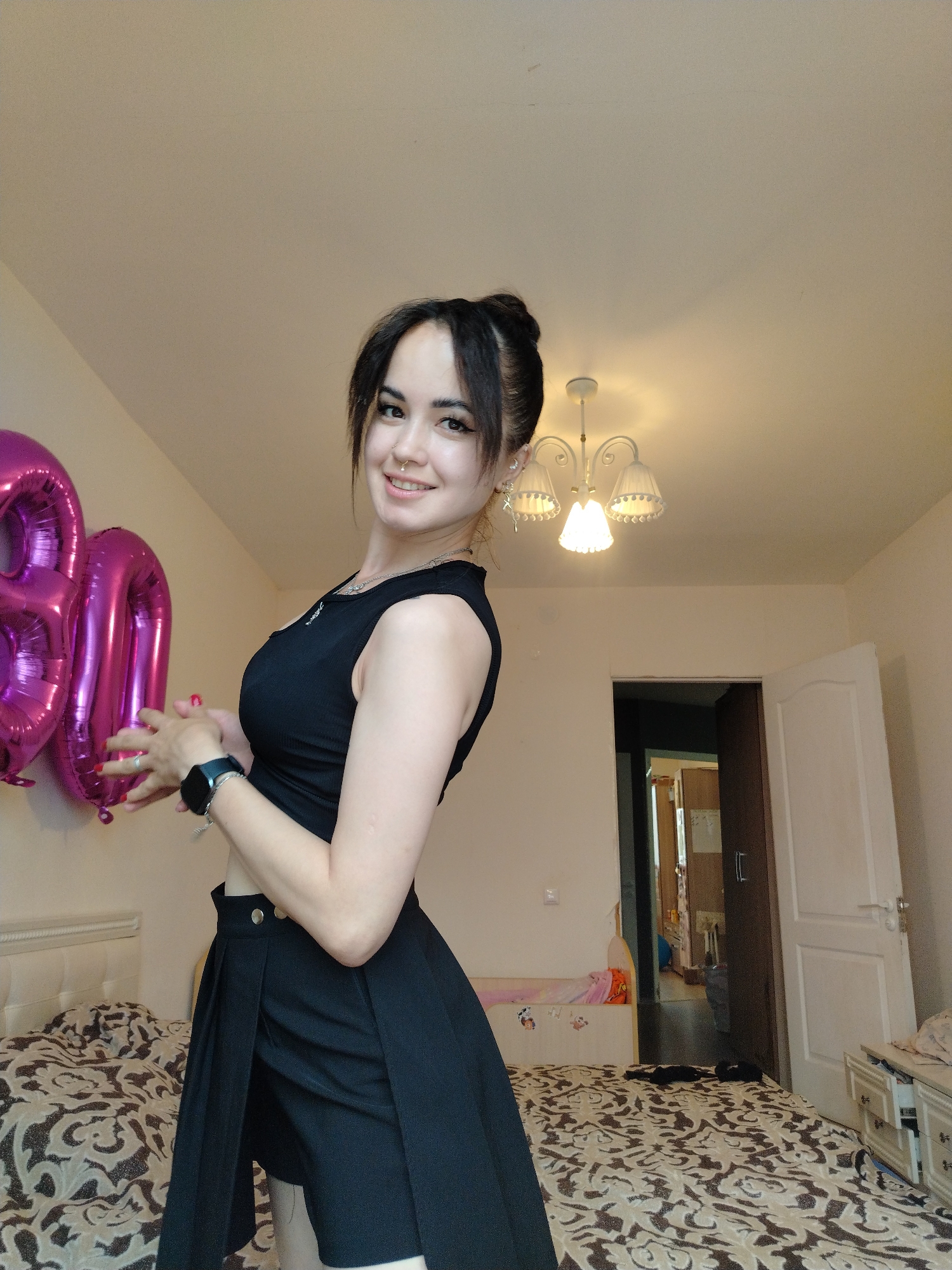 A young woman stands in a bedroom, holding two large, magenta number balloons that appear to say "80". She is wearing a black sleeveless top and a black skirt with shorts underneath. She has dark hair pulled back in a bun and is smiling at the camera. A smartwatch is visible on her left wrist. The room has a bed with a patterned duvet, a white headboard, and a chandelier on the ceiling. A doorway leads to another room, and a white door is slightly ajar on the right. The room is likely in a residential apartment or house. There are no explicit indicators of the city of Noviy, Russia.