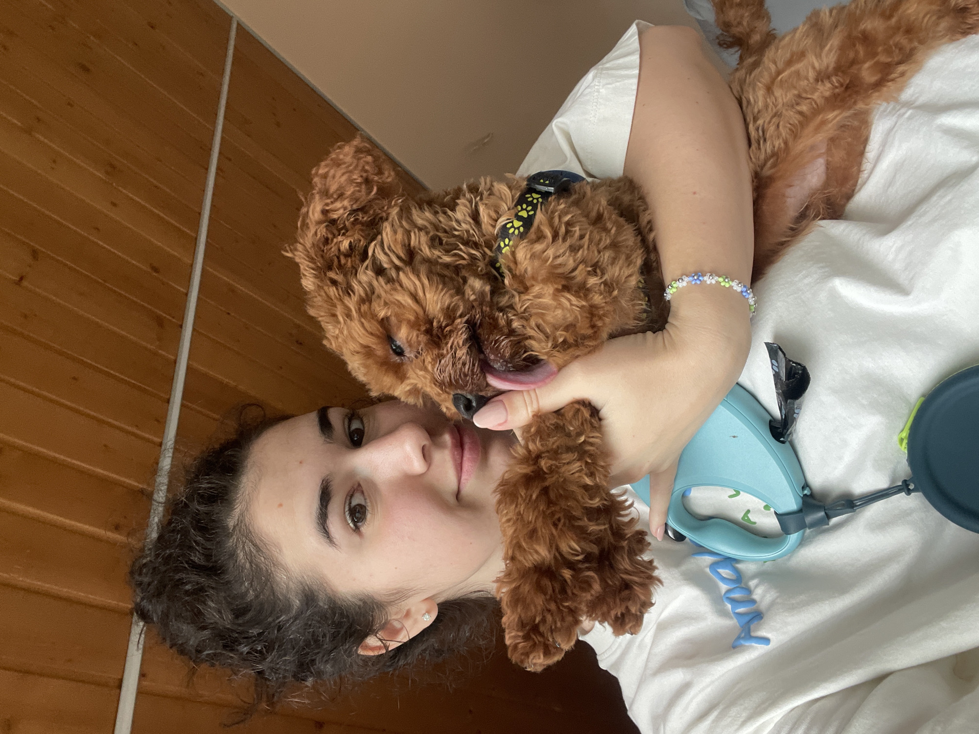 A young woman with dark, curly hair is lying on a white surface, possibly a bed, and embracing a reddish-brown, curly-haired dog. The dog is wearing a black collar with yellow paw prints. The woman has light skin and is wearing a white t-shirt. She is also wearing a beaded bracelet on her right wrist. A light blue dog leash and a dark green dog bowl are visible on the white surface near the woman and the dog. Behind the woman, on the left side of the image, is a wall of light brown, horizontal wooden planks. There is no visible information to determine if the location is in Sofia, Bulgaria.