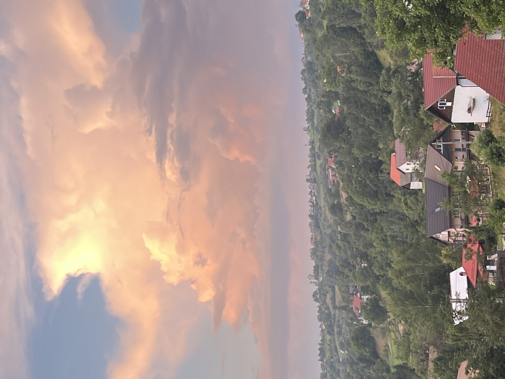 The image depicts a serene sunset over a hilly, rural landscape. A vibrant, peach-colored cloud formation dominates the upper portion of the frame, contrasting with the pale blue sky. Below the sky, a lush green hillside is dotted with numerous houses, many featuring red or brown tiled roofs. The houses are nestled amongst dense foliage, with trees of varying shades of green covering the undulating terrain. In the foreground, several houses are more clearly visible, showcasing architectural details like windows, chimneys, and balconies. The overall impression is one of tranquility and natural beauty, with the warm hues of the sunset casting a peaceful glow over the scene.