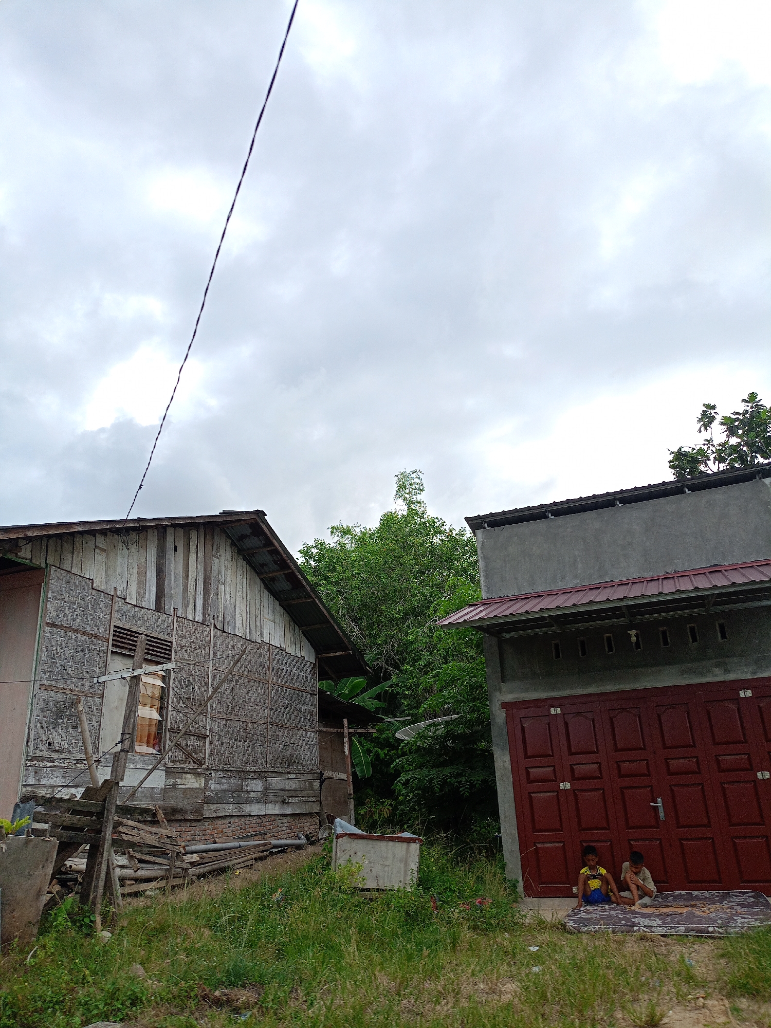 Two young children are sitting on a mattress in front of a dark red double door in Meunasahteungkudigadong, Indonesia. The door is part of a gray building with a dark red roof overhang. To the left is an older, weathered wooden building with a woven exterior. Green vegetation and trees surround the buildings. An electrical wire stretches across the sky, which is overcast with gray clouds. Debris and overgrown grass fill the foreground.
