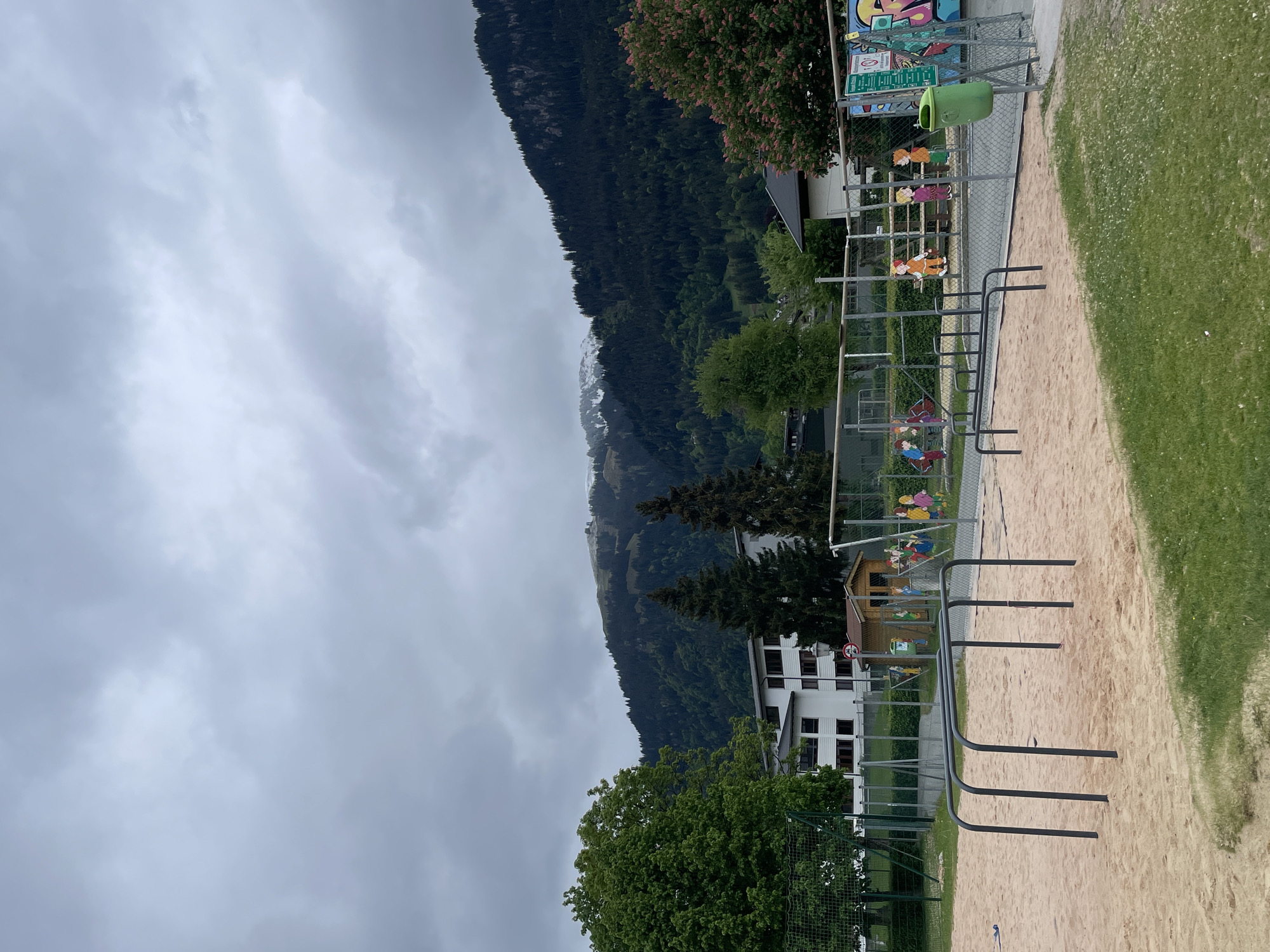 The image depicts a playground located in an area with mountainous terrain. The foreground features a sand pit with several metal bars. A grassy area is adjacent to the sand pit. A metal fence separates the playground from the surrounding area, with colorful decorations attached to it. In the background, a building with white walls is visible, partially obscured by trees. Beyond the building, a mountain range covered in dense forest rises against a cloudy sky. A small area of snow is visible at the higher elevations of the mountains. The sky is overcast with grey clouds.