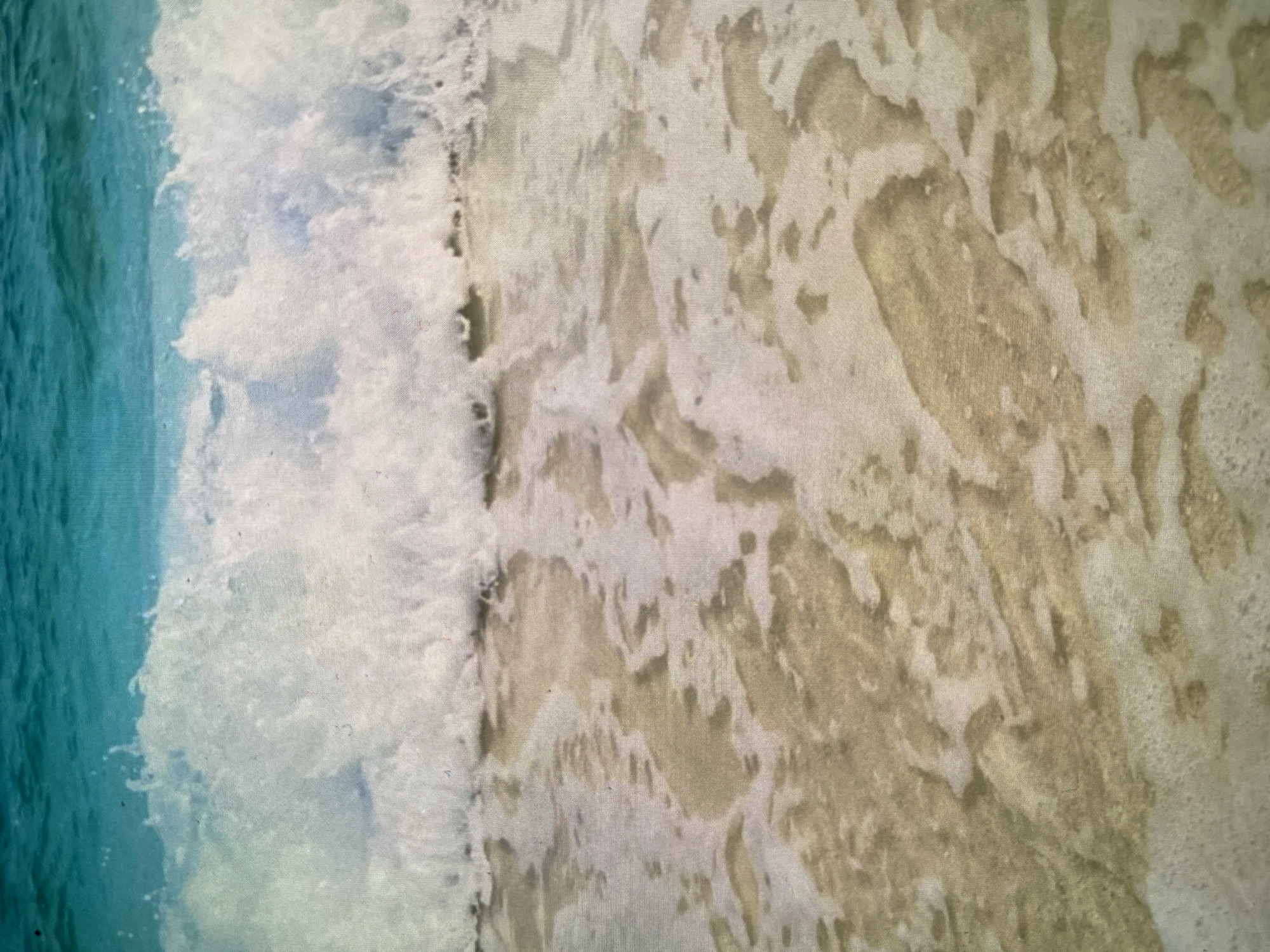 The image captures a close-up, overhead view of ocean waves breaking on a sandy shore. On the left side, the vibrant turquoise-blue water of the ocean transitions into a dynamic mass of white foam as a wave washes towards the right. This foamy wave front creates intricate, swirling patterns on the light-colored sand.
As the wave recedes, it leaves behind a mosaic of wet sand and residual foam, forming abstract shapes and textures across the majority of the frame. The sand appears damp and light beige, contrasting with the brighter white of the foam. The overall impression is one of movement and natural beauty at the waterline.
There are no people, man-made objects, or visible text in the frame. The lighting suggests bright daylight conditions, and the scene conveys a serene, natural beach environment. The image quality reveals a slightly pixelated or textured appearance, possibly due to it being a photograph of a screen or print, which gives the surface a subtle grid-like pattern. The location appears to be a generic sandy beach, without specific cues to identify its precise geographic location as Bankya, Bulgaria.
