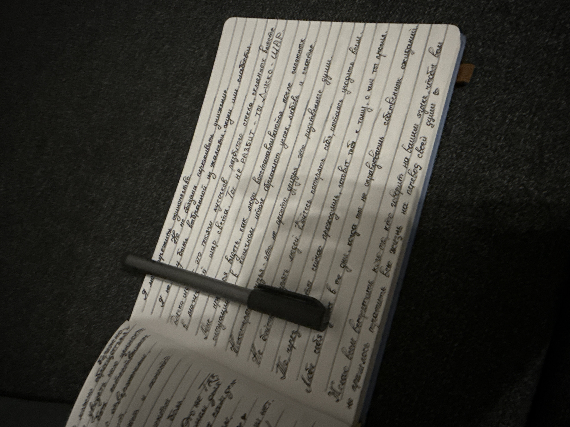 Here's a detailed description of the image:
**General Description:**
The image shows a notebook open on a dark surface. A pen lies across the open pages. The notebook appears to be filled with handwritten text in the Russian language. The handwriting is cursive.
**Objects:**
* **Notebook:** A lined notebook with rounded corners.
* **Pen:** A dark-colored pen with a clip.
* **Text:** Russian cursive handwriting covering the pages.
**Environment:**
* The background is a dark, somewhat textured surface.
**Interpretation and Subject:**
The notebook appears to contain personal writings, likely a diary, journal, or creative writing piece, based on the dense text. The placement of the pen suggests active use of the notebook.
There is no way of telling whether the location is Saint Petersburg, Russia, without additional contextual information from the picture.