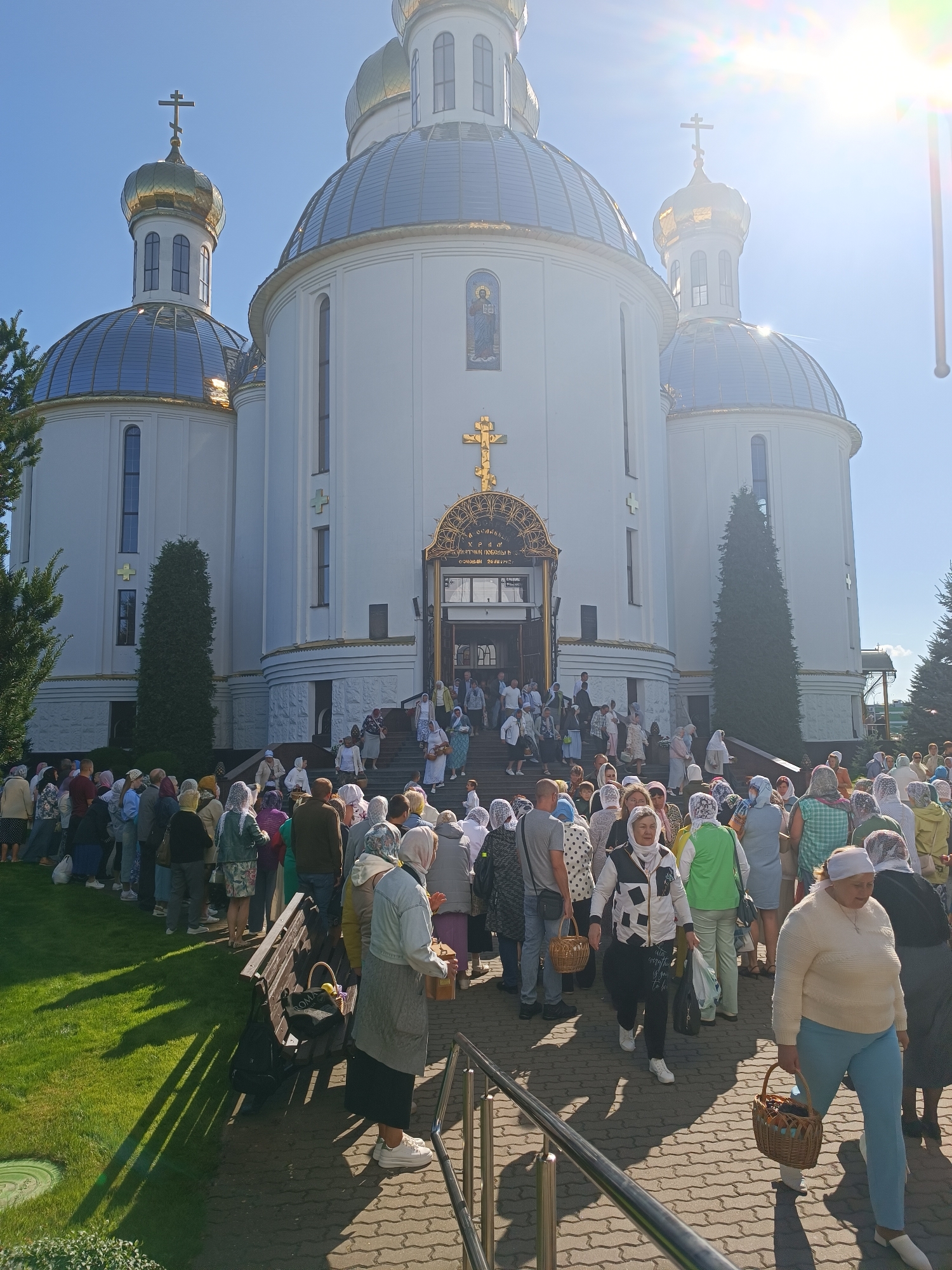 A crowd of people, mostly women wearing headscarves, are gathered outside a large white church with golden domes. The church has a grand entrance with an arched doorway, and a golden cross above it. The people are standing in a queue, some are walking up the stairs towards the entrance, and others are standing on the grass. Many are carrying baskets. The sun is shining brightly, casting long shadows on the ground. The scene appears to be taking place in the city of Brest, Belarus, based on the context provided.