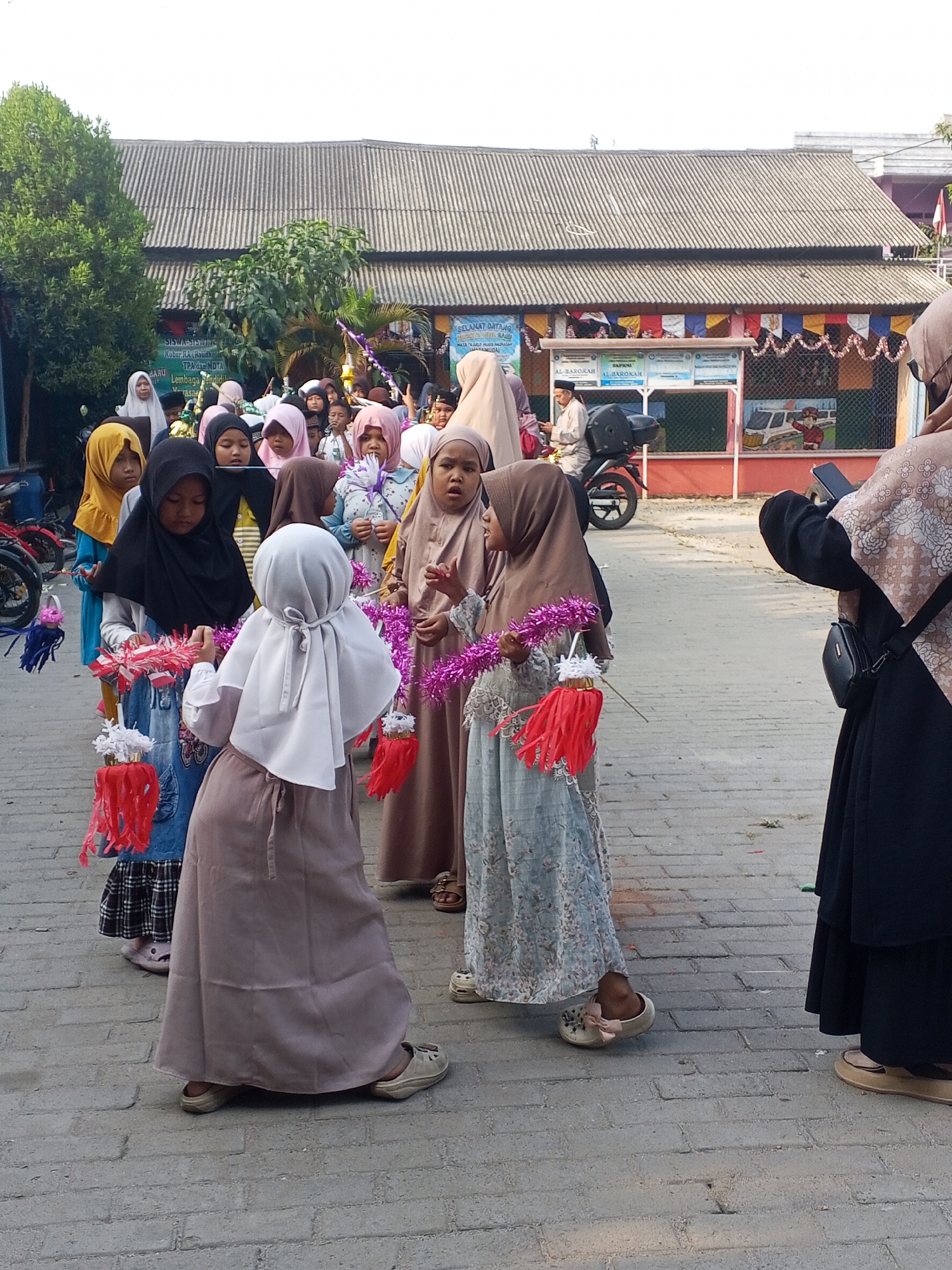 The image shows a group of young girls in modest Islamic clothing, likely students from a school in Ciruas, Indonesia, participating in an outdoor activity or parade. They are dressed in hijabs and long dresses, some holding decorative items such as fringed paper lanterns and tinsel. The setting appears to be a schoolyard or paved outdoor area in front of a building with a corrugated metal roof. The background shows a building with colorful banners and murals, and some foliage. The overall atmosphere suggests a celebratory or educational event for children.