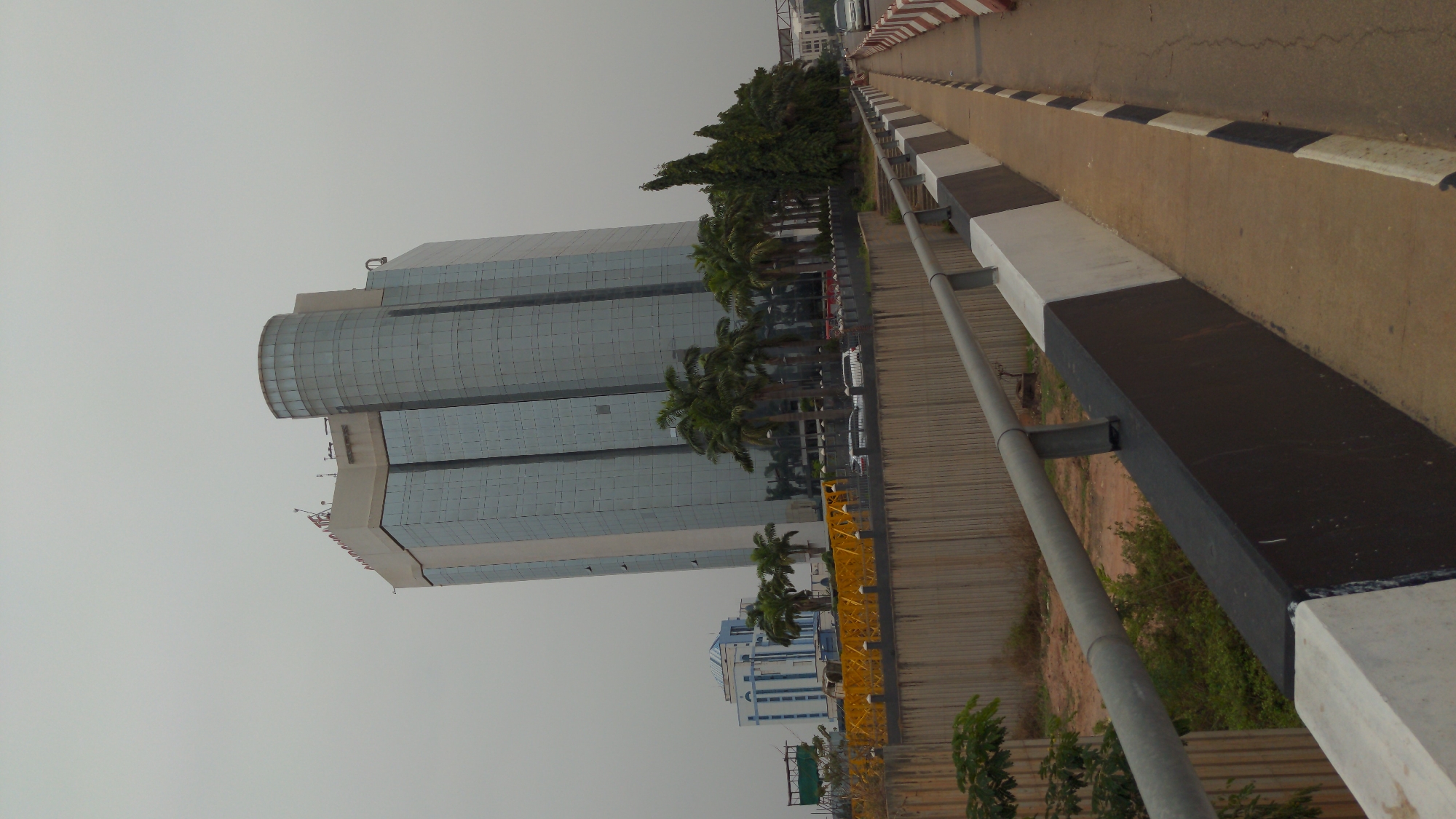This image captures a modern, glass-clad skyscraper dominating the left side of the frame, under an overcast sky. The tall building has a distinctive design, featuring a prominent, rounded, glass-enclosed section near its top. Faint text, possibly a company name such as "NNPC" or "NNPC Towers," is visible on the upper part of the building, consistent with its location in Abuja, Nigeria.
In the foreground on the right, an elevated concrete pathway or bridge extends into the distance. This path is clearly defined by black and white painted concrete barriers and an accompanying grey metal guardrail. Below the pathway, the ground is uneven, showing dirt and sparse green vegetation.
Between the main building and the elevated path, several palm trees and other green foliage are visible, partially obscuring the lower sections of the skyscraper and a few parked or moving vehicles. A smaller, multi-storied building with blue and white exterior details is also visible further back and lower down. The overall scene depicts an urban or semi-urban environment during daytime, characterized by diffused light from an overcast sky. No specific human activity or event is taking place.