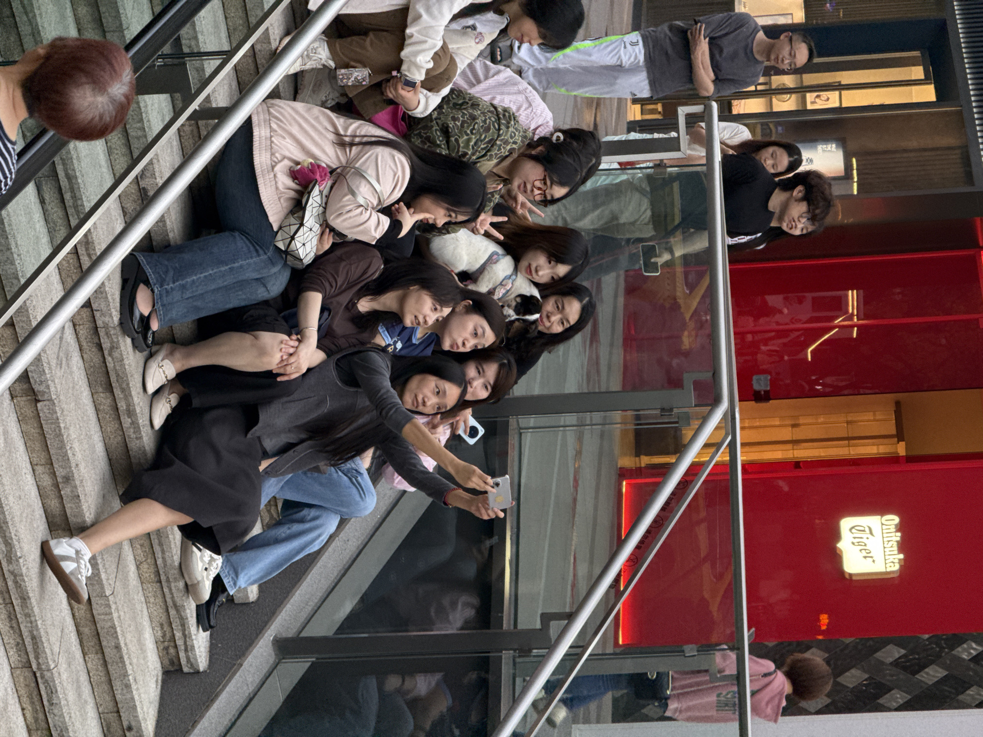 A group of approximately nine young women are sitting on outdoor stairs, posing for a selfie. One of the women holds a phone to take the photo. They are positioned on the steps with their heads close together, facing towards the camera. Other individuals are visible in the background on the street. A red sign for "Onitsuka Tiger" is visible in the background. The location is Yuzhong District, Chongqing, China, based on the sign and architectural style.