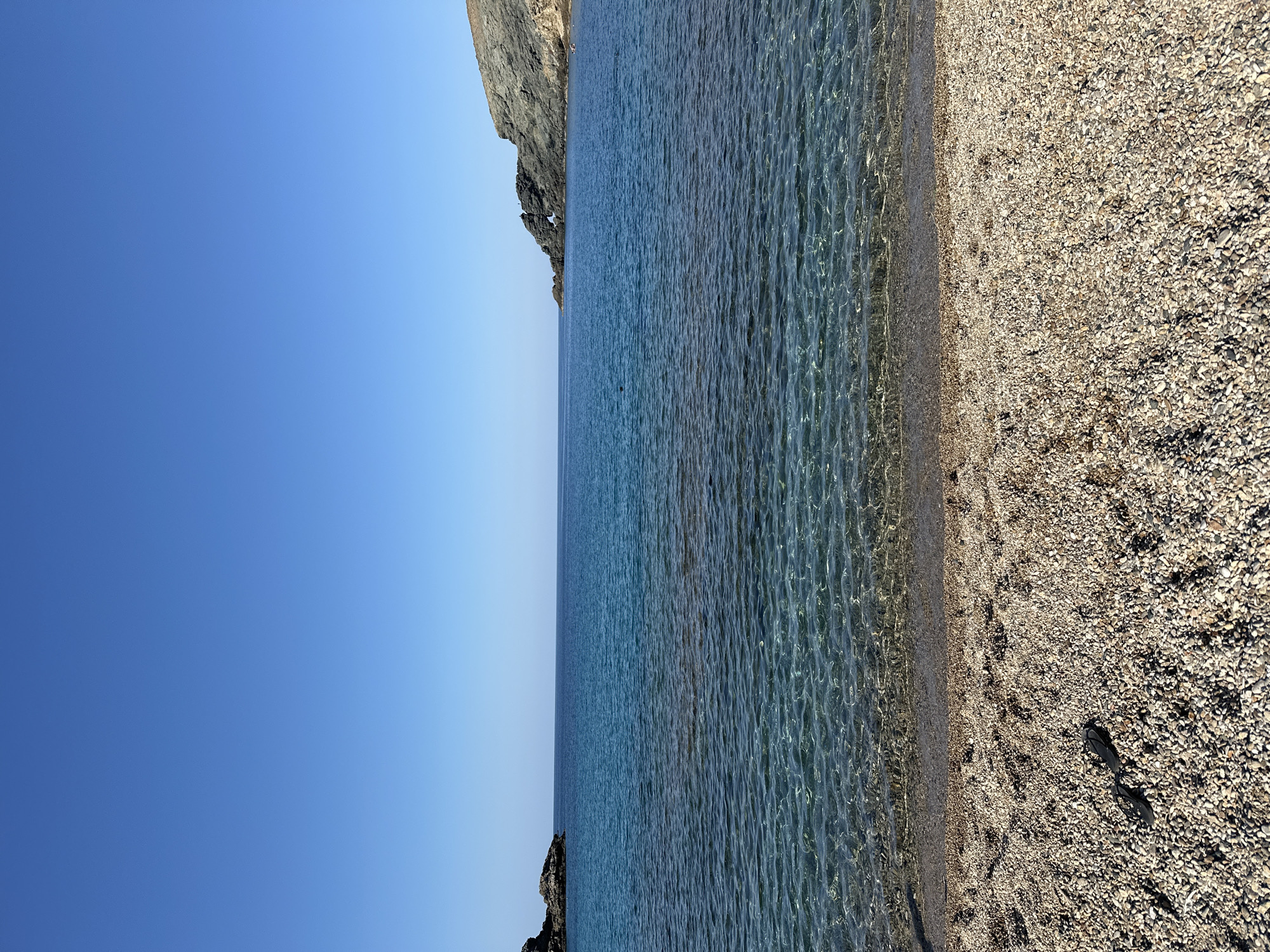 The image depicts a serene beach scene in Ano Mera, Greece, under a clear, bright blue sky. The foreground is a pebble beach with a pair of black flip-flops lying on the pebbles. The water is crystal clear, revealing the pebbles on the seabed near the shore. The water transitions to a deeper blue further out, with gentle ripples on its surface. In the mid-ground, a rocky outcrop juts out into the sea on the right, and a smaller rocky formation is visible on the left. The horizon is sharp, separating the sea from the sky. Two small figures are visible swimming in the water. The overall scene conveys a sense of peace and natural beauty.