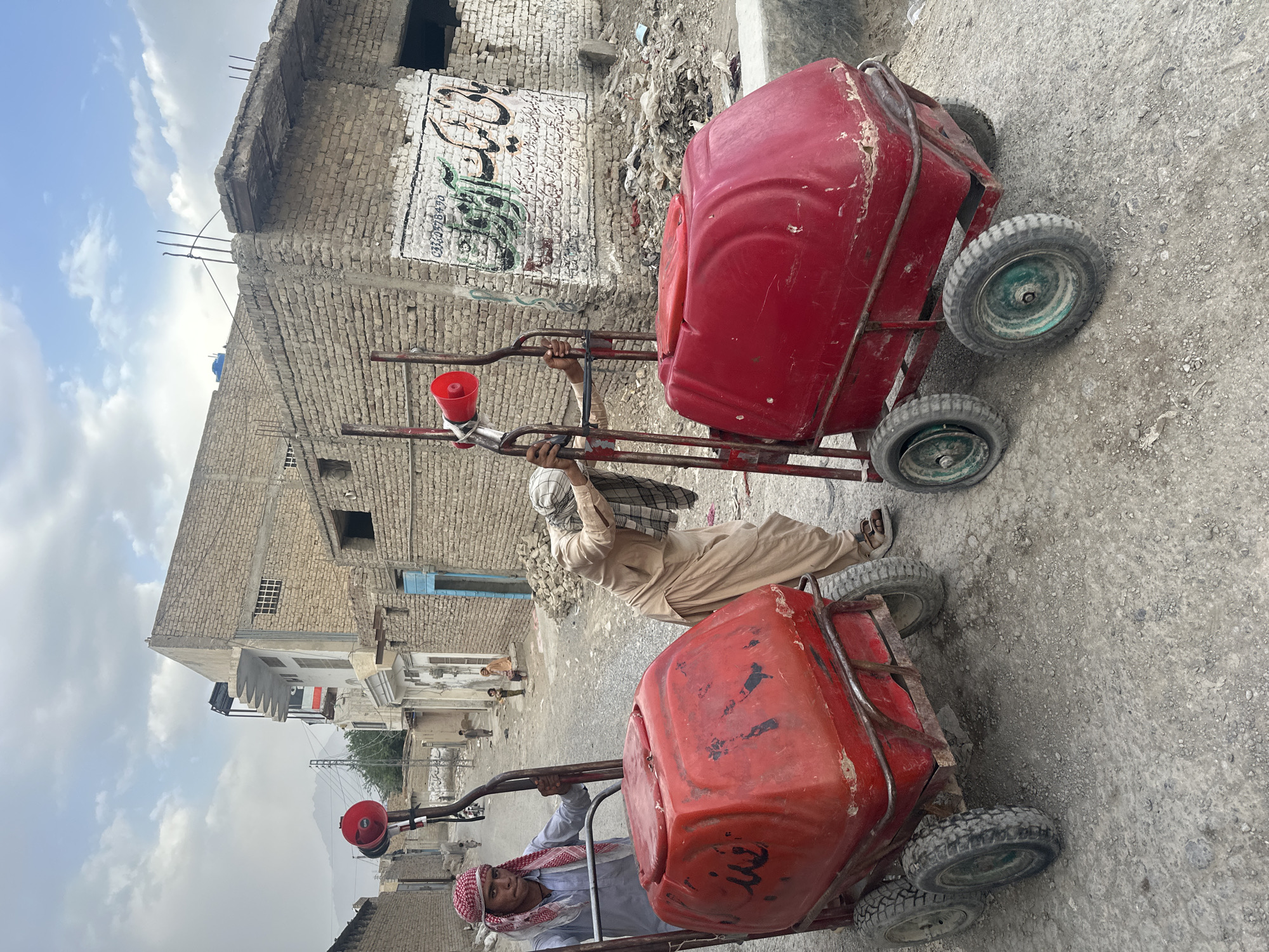 The photograph depicts two individuals in Quetta City Tehsil, Pakistan, each pulling a red cart on a rough, unpaved street. The carts appear to be carrying red containers. The individuals are dressed in traditional Pakistani clothing, including head coverings. The background features buildings with weathered brick facades and graffiti. The sky is overcast with patches of blue.