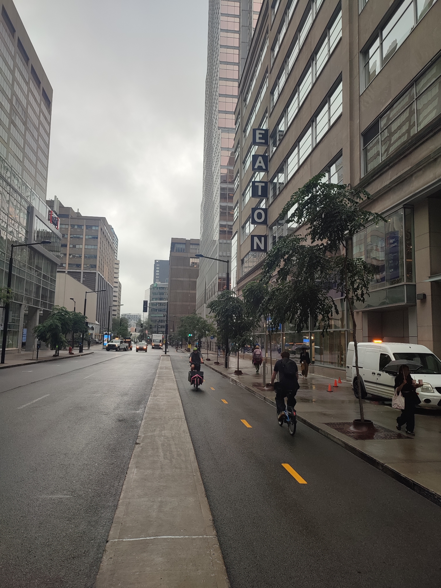 The image depicts a street scene in Montreal, Canada, under an overcast sky. The perspective is from the sidewalk looking down a wet street lined with tall buildings. On the right side of the street, there is a building with a prominent sign that spells out "EATON" vertically. Trees are planted along the sidewalk in front of this building. A white van with an orange flashing light is parked on the right. Further down the street, several cars are visible, including a white truck. On the left side of the street, there are more buildings and trees. A cyclist is riding on a dedicated bike lane marked with yellow lines, and another cyclist is further ahead. A person is walking on the sidewalk with an umbrella, and another person is walking behind them. The street appears wet, suggesting it might be raining or has recently rained.