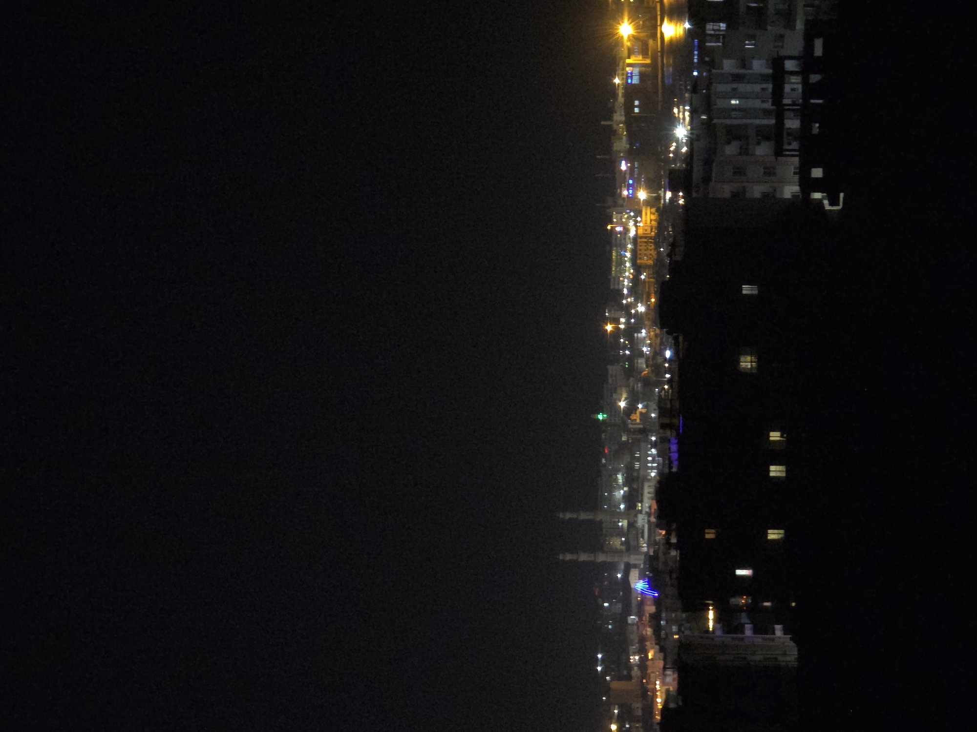 The image depicts a nighttime view of a city skyline along a body of water, likely a coastline or harbor. The scene is dominated by the dark night sky and the illuminated cityscape. Tall buildings, some appearing to be residential or commercial, are visible with numerous windows lit up, creating a pattern of yellow and white lights. Streetlights and other artificial lights line the waterfront, casting a warm glow. In the distance, there are structures that resemble minarets, suggesting a location with Islamic architecture, consistent with the Al Hadaba, Egypt context.
The activity implied is the normal functioning of a city at night, with lights indicating occupied buildings and ongoing activity. The water in the foreground reflects some of the city lights, creating a shimmering effect. The overall mood is quiet and serene, characteristic of a city at rest. The time of day is clearly night, as indicated by the dark sky and artificial lighting. There are no discernible people or specific objects that are the primary focus, but rather the collective presence of the urban environment. No visible text is present in the image. The composition suggests an elevated viewpoint, looking down upon the city and water.