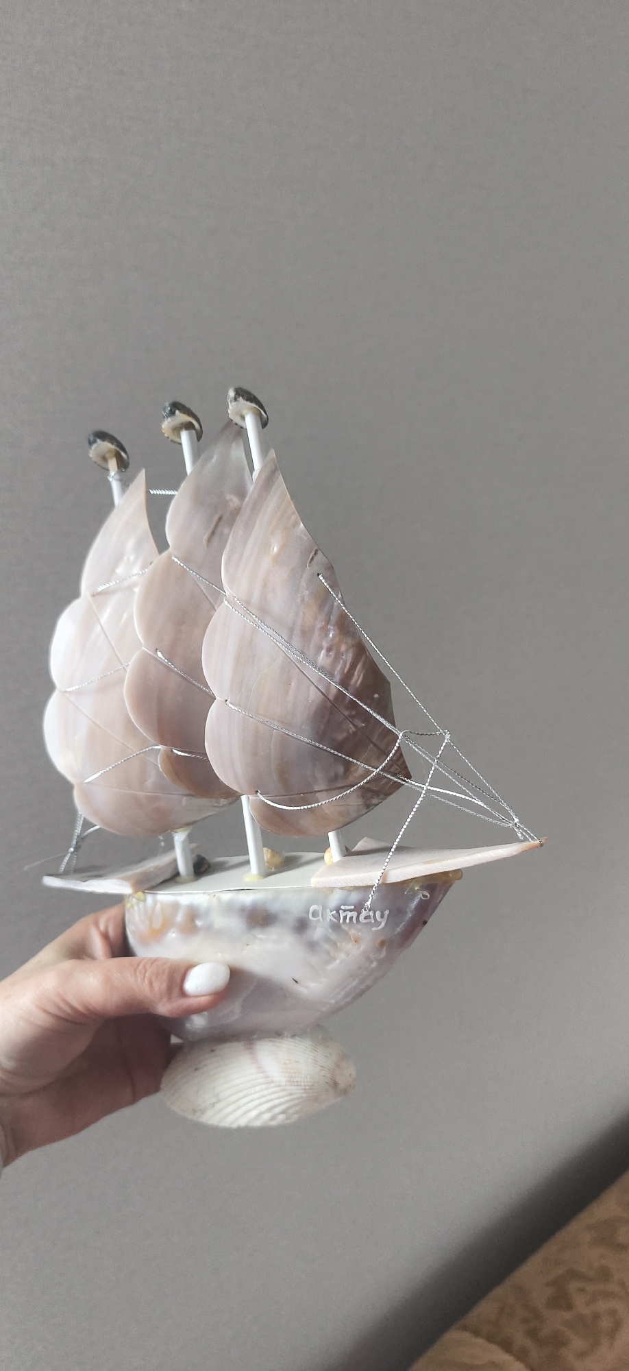 A hand holds a decorative sailboat model against a solid gray background. The boat's hull and base are constructed from seashells. The sails, also made of shells, are held in place by white poles and silver string. The name "Akmay" is written on the side of the boat. The location is Фоки, Russia.