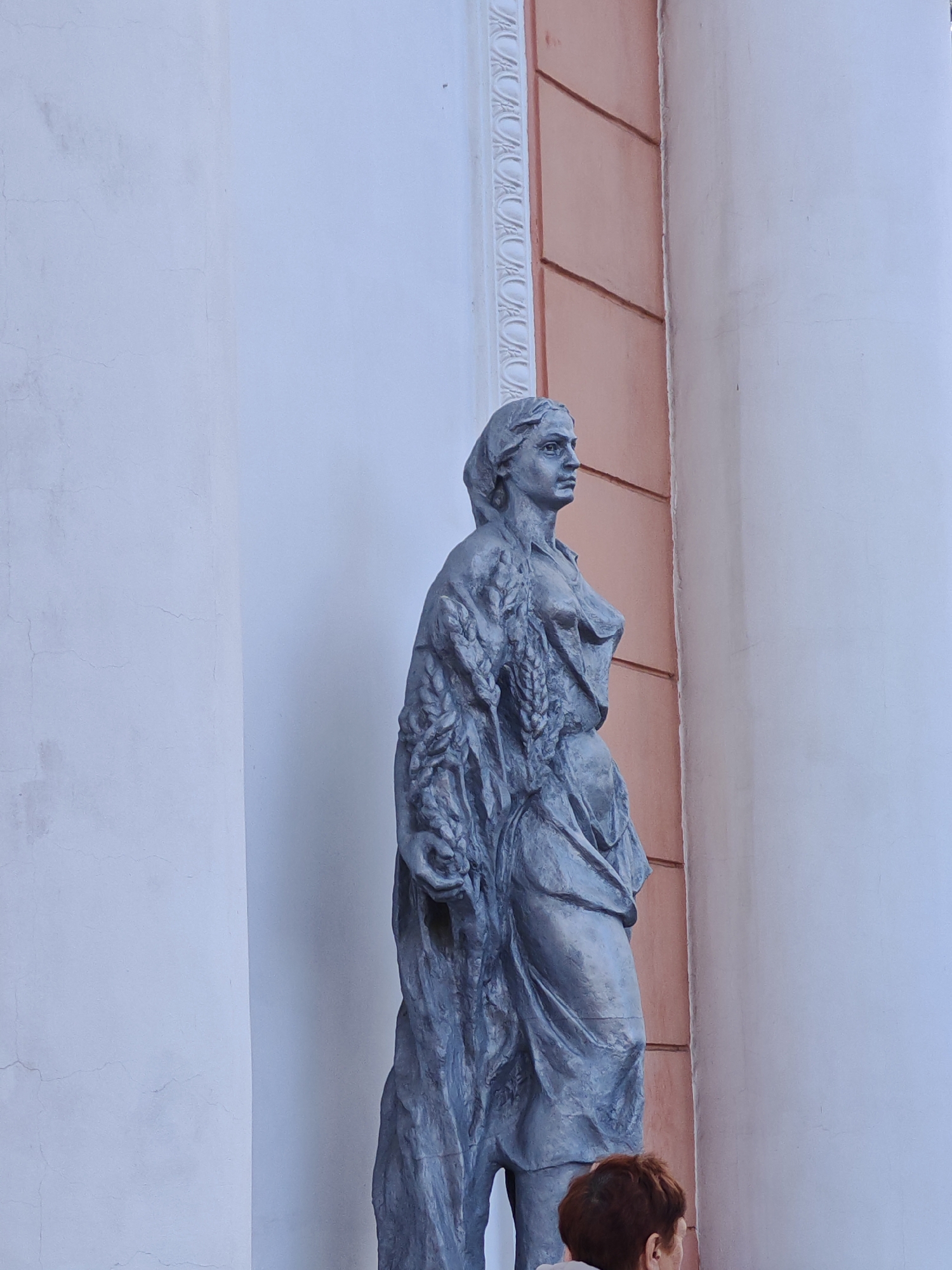 The image shows a stone statue of a woman holding wheat stalks, in front of a building with white and peach-colored columns. The statue is gray and detailed, with the woman's hair styled in a bun and her clothing draped loosely. The background building features classical architectural elements, including fluted columns and decorative molding. The lighting suggests it is daytime. A person with short, reddish-brown hair is visible in the lower right corner of the frame, looking towards the statue. The statue appears to be located in an outdoor public space, possibly a park or square, in the city of Homyel, Belarus.